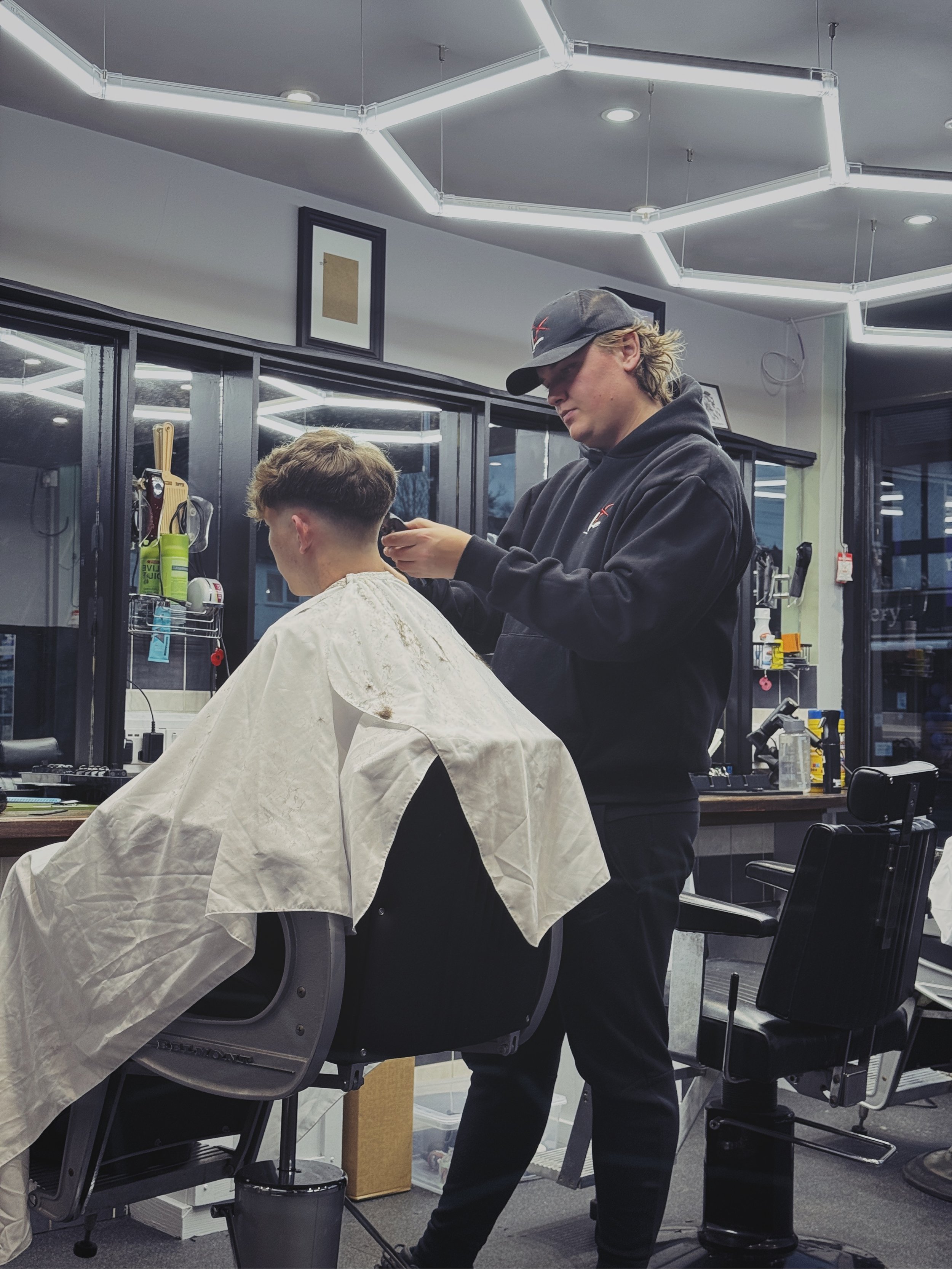 Barber cutting a young man's hair in a modern barbershop with black chairs and large windows.