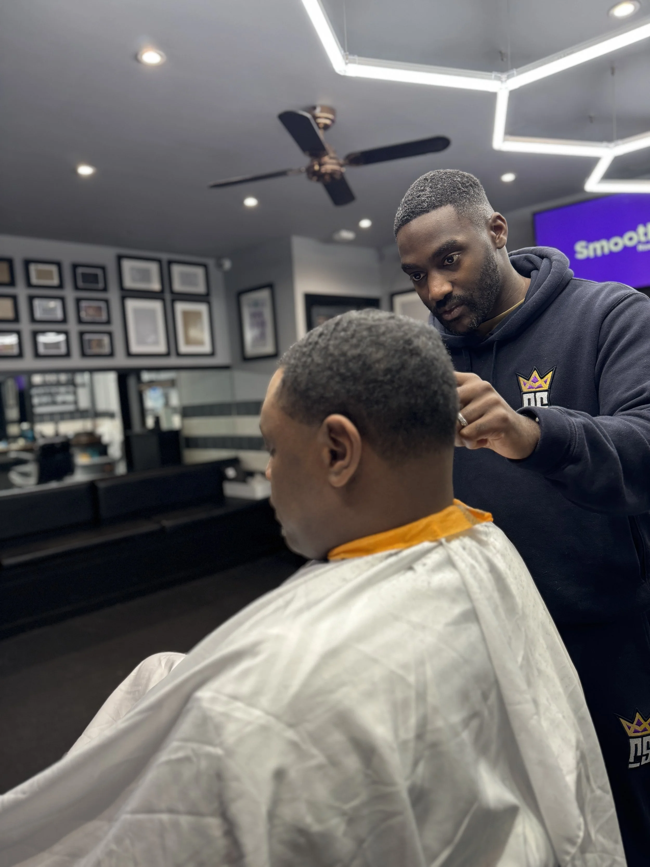 Barber giving a haircut to a man in a modern barbershop with framed pictures on the wall and a ceiling fan.
