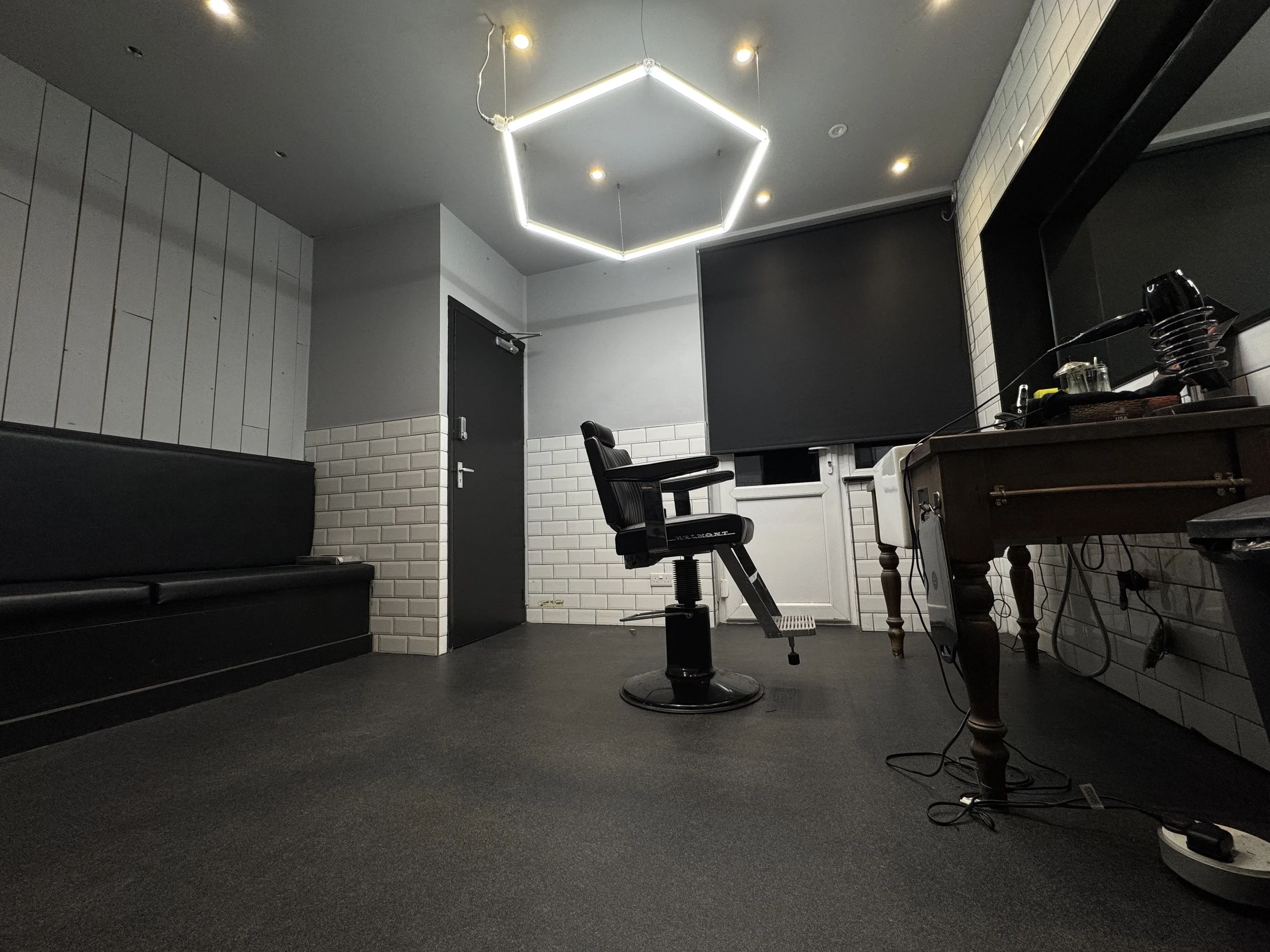 Empty room with black leather bench, black swivel chair, white brick walls, black roller blind on window, dark grey ceiling with modern hexagonal light fixture, and wooden desk with various items on it.