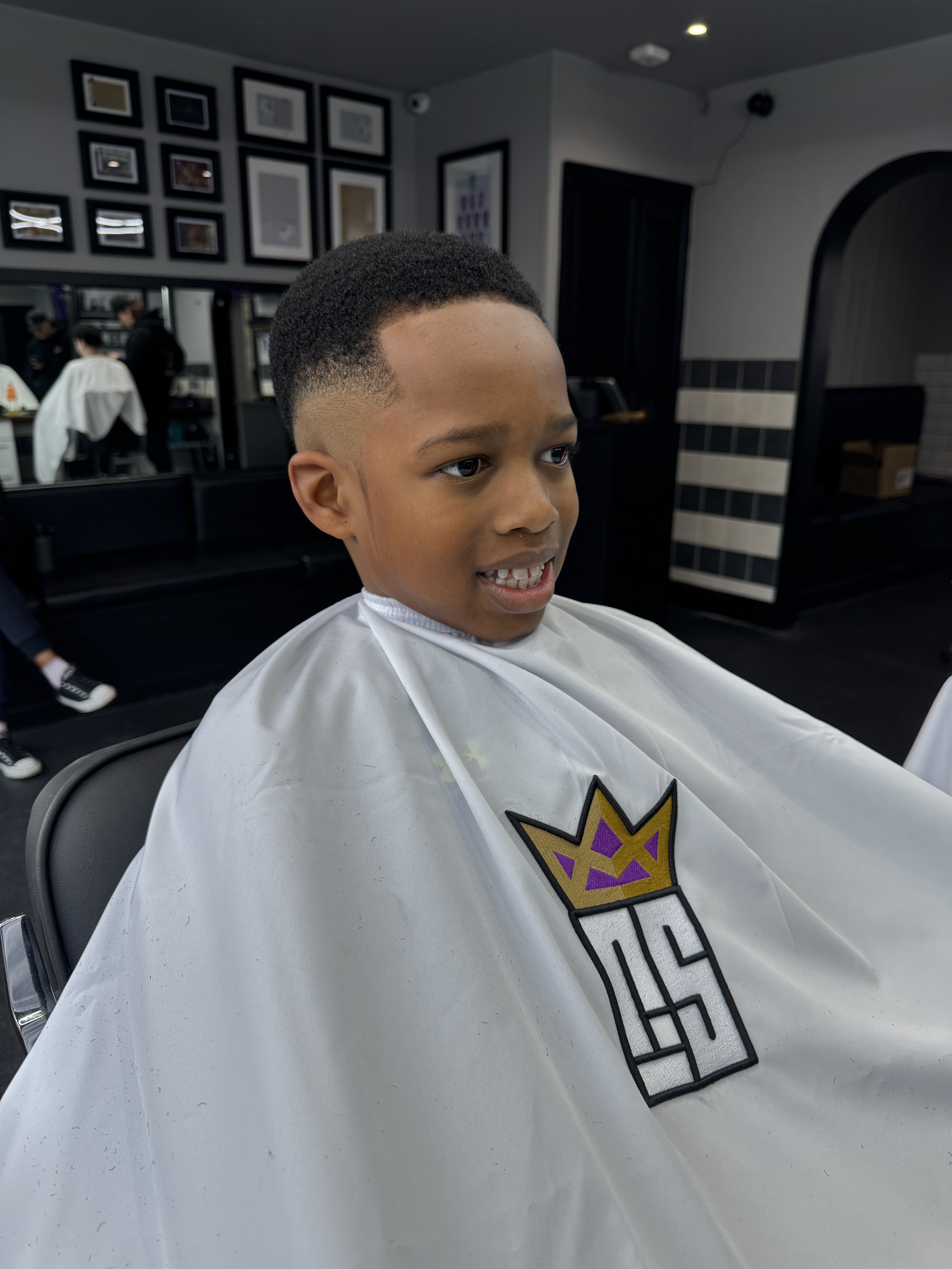 Young boy with a fresh haircut sitting in a barber shop, wearing a barber cape with a logo of a crown and the number 54.