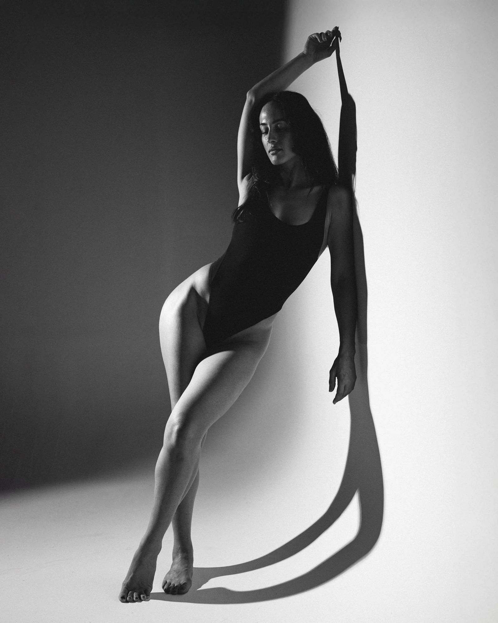 Black and white photo of a woman with long hair in a leotard, posed with one arm raised holding a ribbon, casting a shadow on the wall behind her.