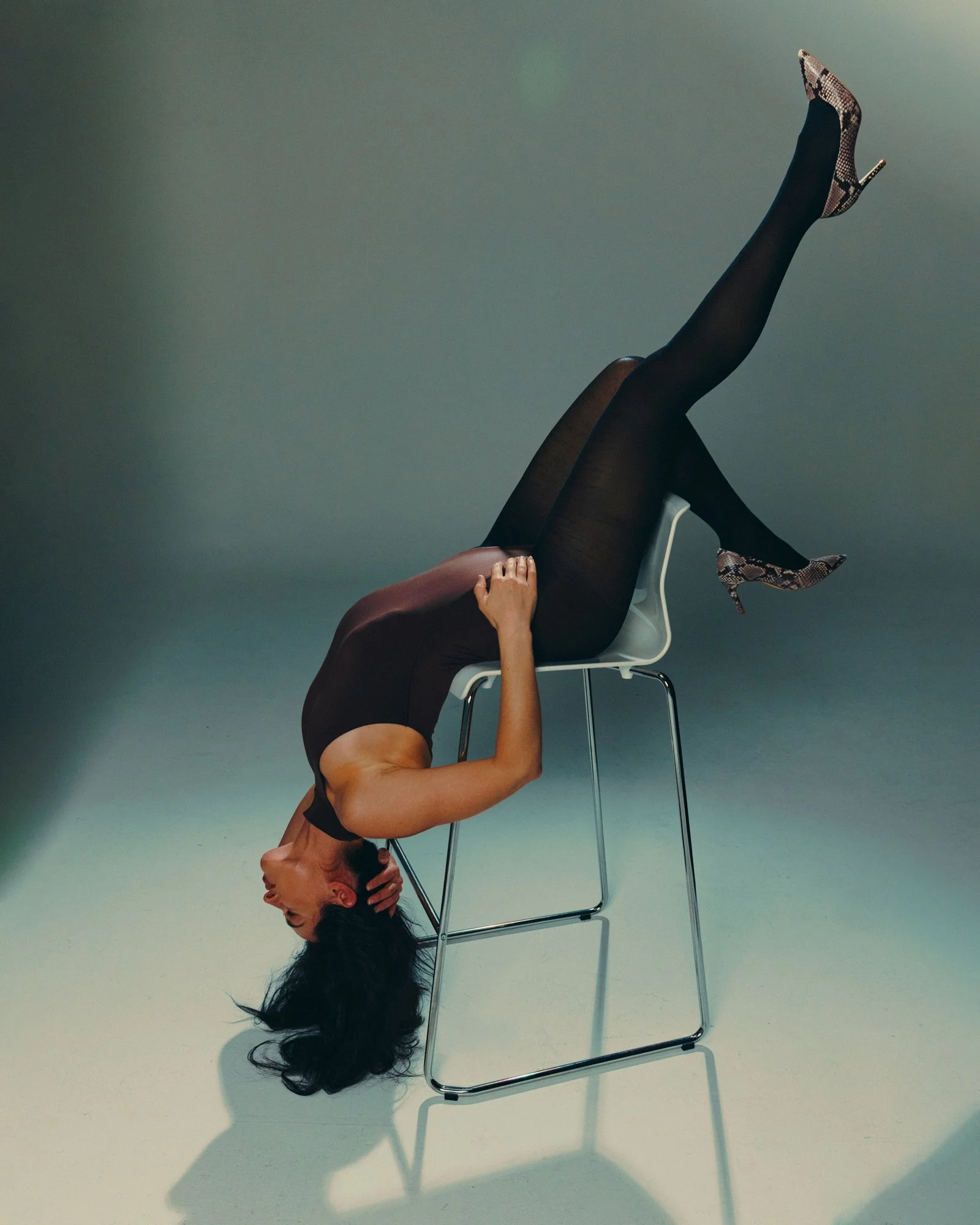 A woman sitting in a chair with her head upside down, legs extended on the chair, wearing black tights and high heels, with hair hanging down and one hand on her head.