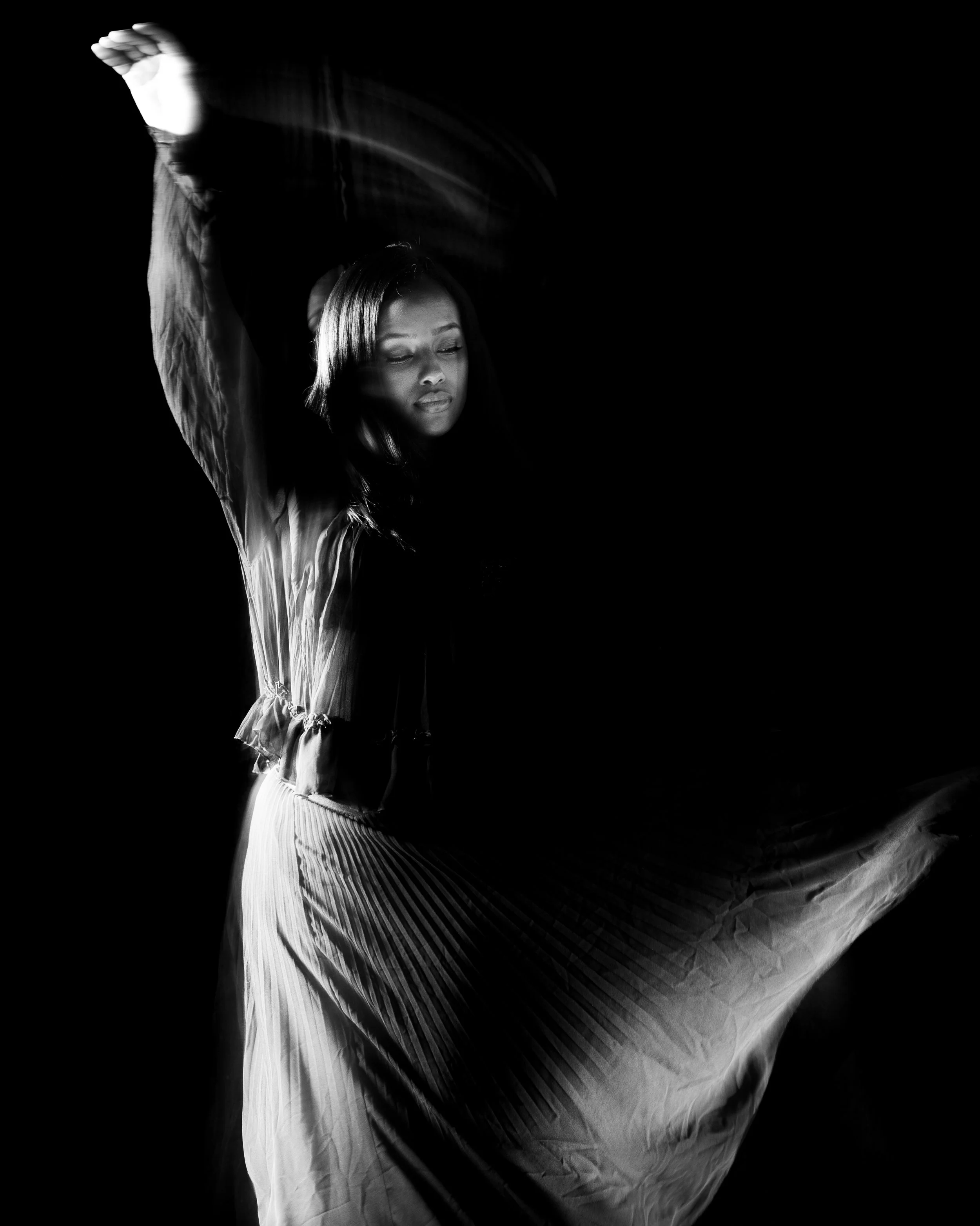 A woman with long hair, closed eyes, and a serene expression, appears to be in motion, with her arm raised above her head, wearing a flowing dress, set against a stark black background.