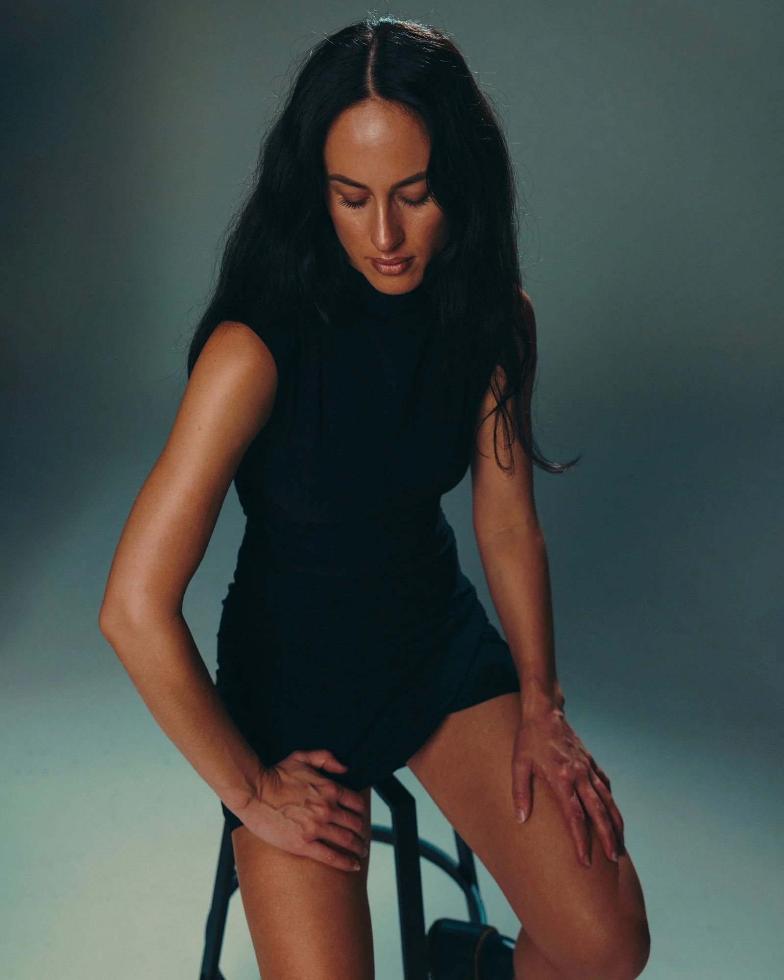 A woman with long dark hair wearing a black sleeveless top, sitting on a black chair, with her eyes closed and head tilted downward, hands on her thighs, against a plain gradient background.