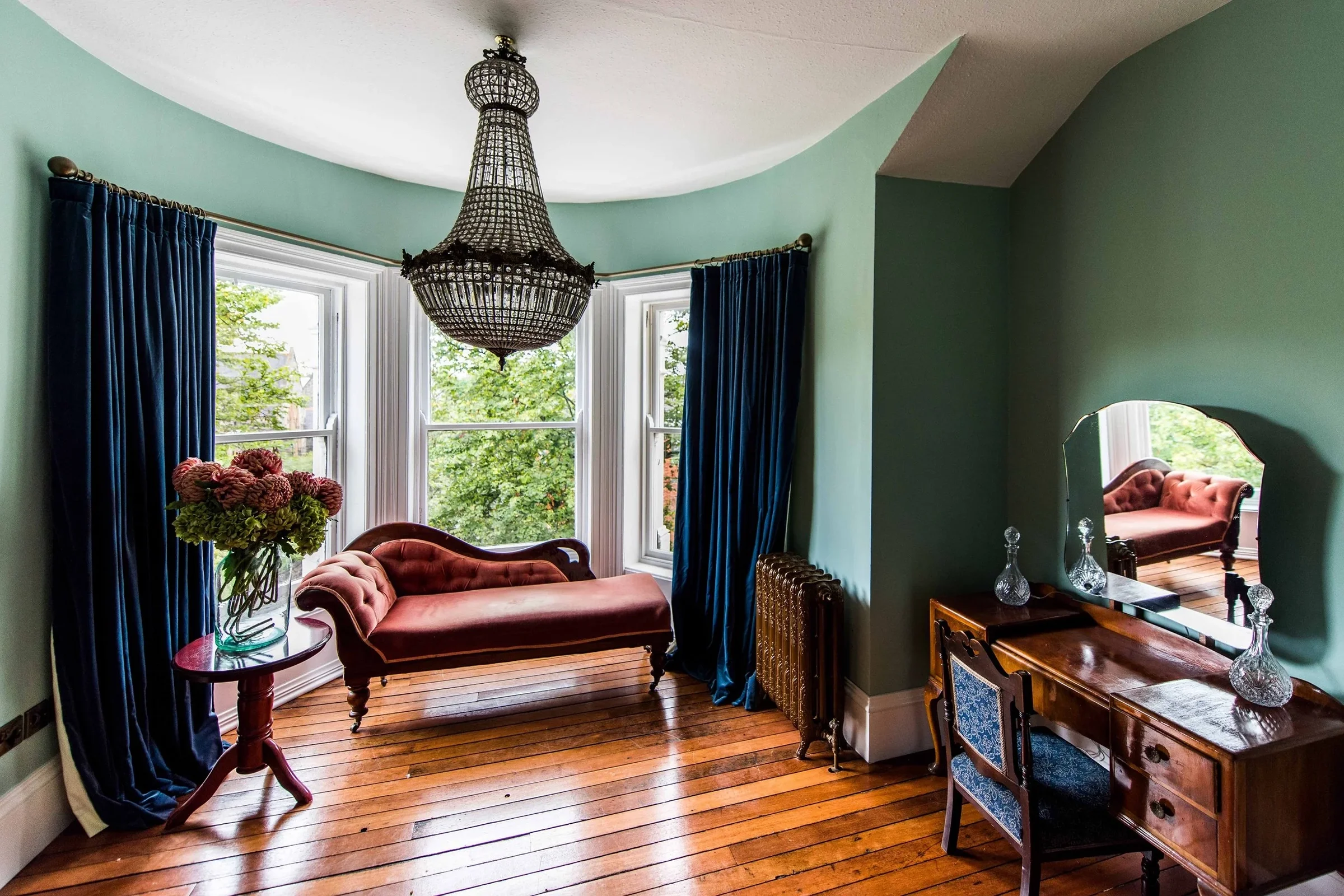 A vintage-style sitting room with teal walls, a large window with dark blue curtains, a pink velvet settee, a wooden vanity with a mirror, glass decanters, a floral arrangement in a glass vase, and a brass radiator. A black chandelier hangs from the 