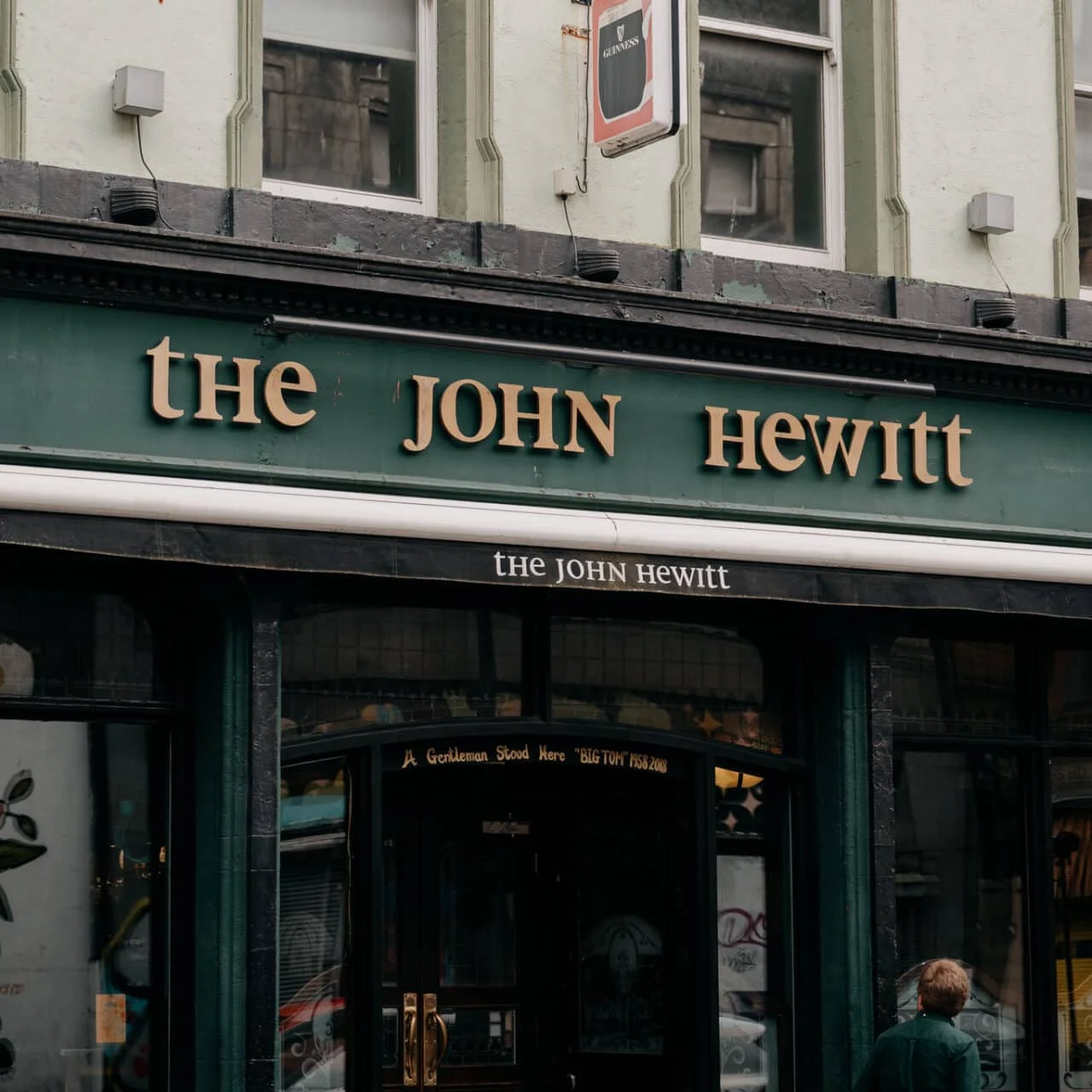 The exterior of a pub called 'The John Hewitt' with green signage, gold lettering, and a black awning. A person with brown hair, wearing a green jacket, is standing outside.
