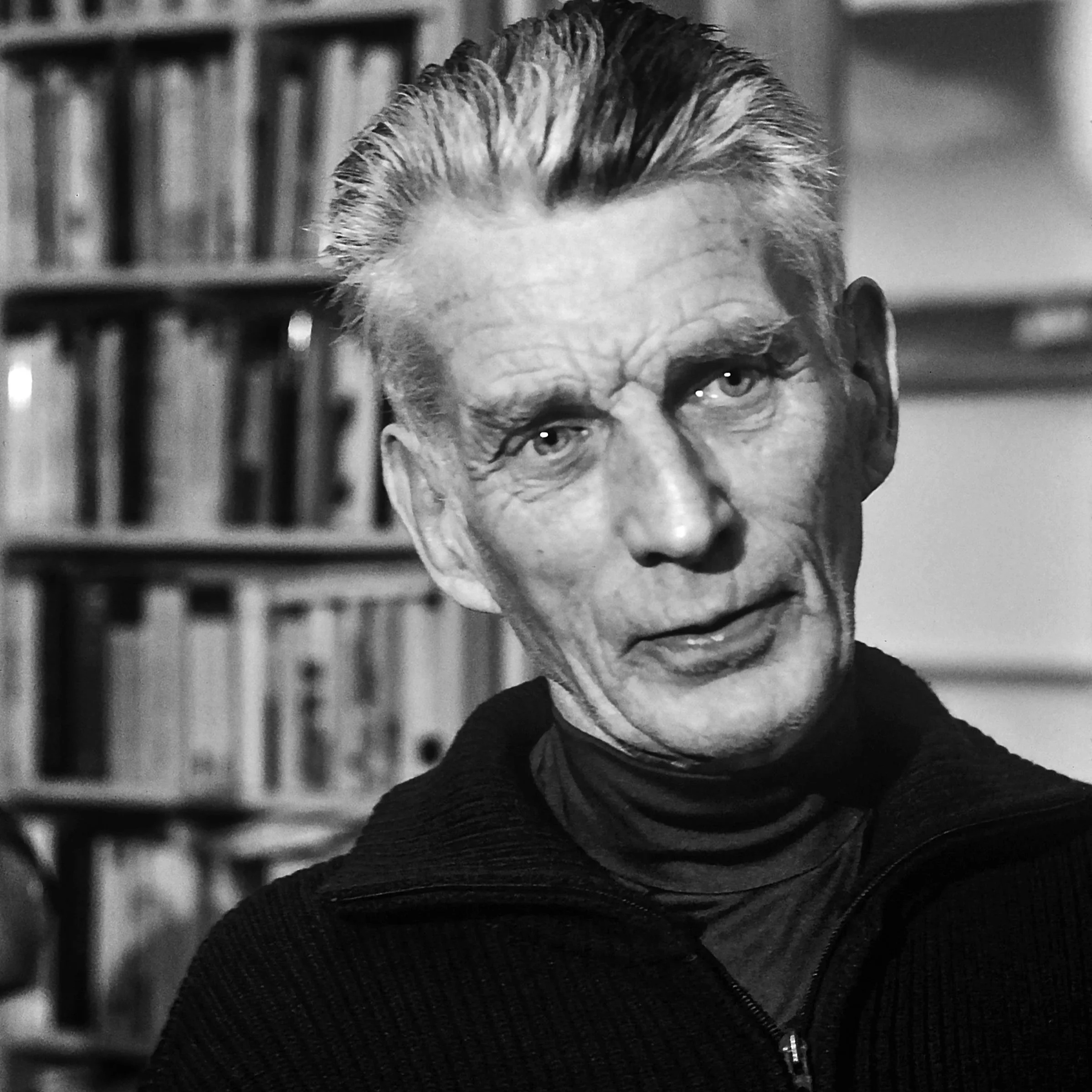 Black and white photo of an older man with slicked-back hair, wearing a dark turtleneck and zip-up sweater, looking directly at the camera in front of a bookshelf filled with books.