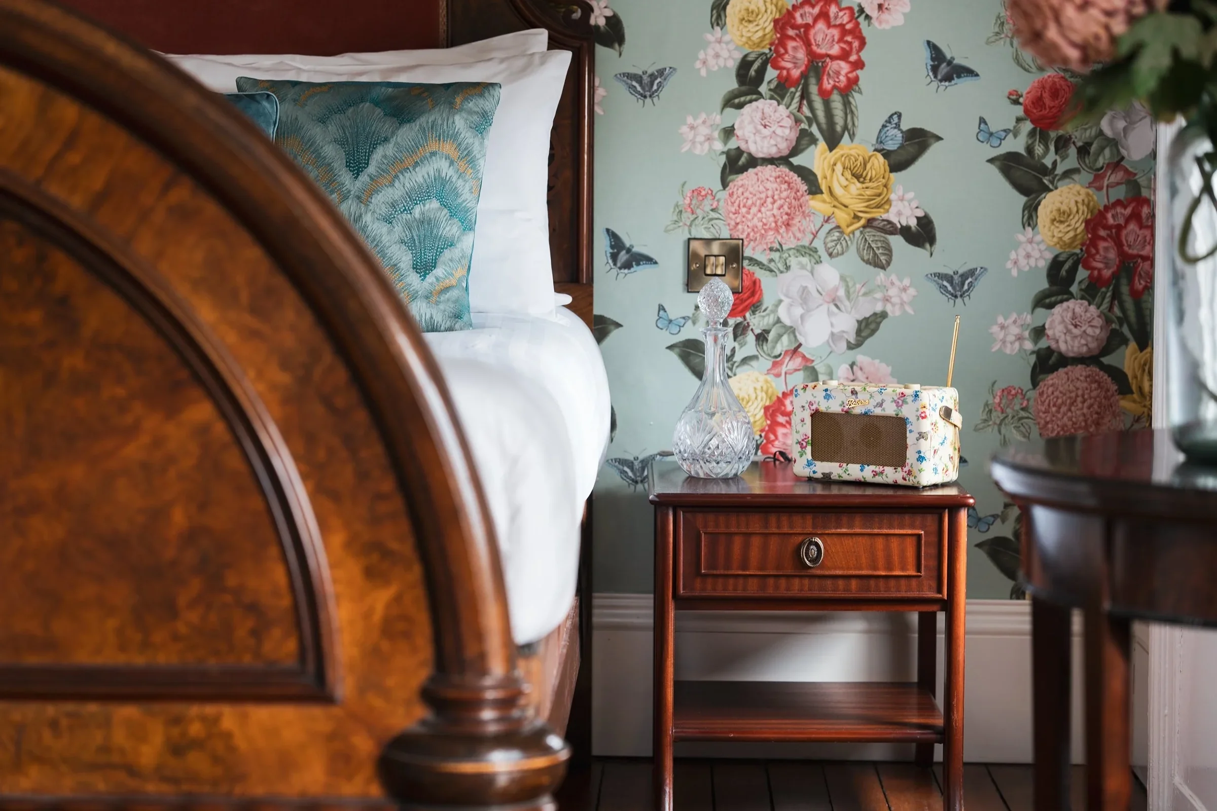 Close-up view of a bedroom corner with a wooden bed frame, white bed linens, a decorative pillow, a dark wood nightstand, a glass vase, a vintage radio, and floral wallpaper with butterflies.