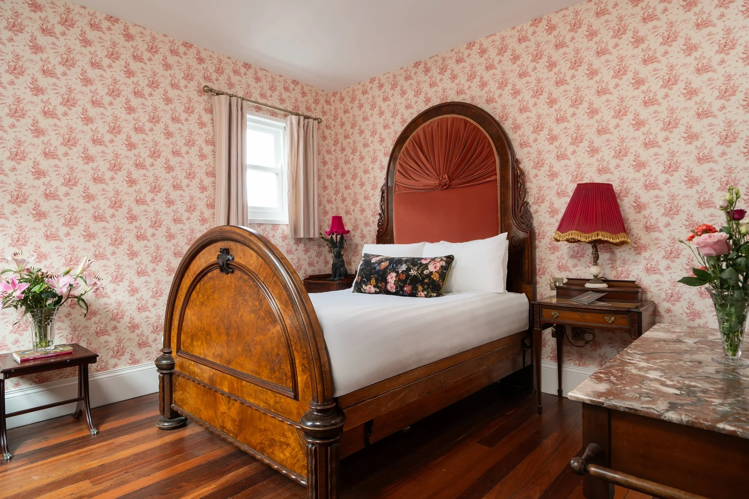 A bedroom with Victorian-style wooden bed frame and floral wallpaper, featuring a small window with curtains, side tables with pink lamps, vase with flowers on a marble-topped dresser, and white bedding with a decorative pillow.