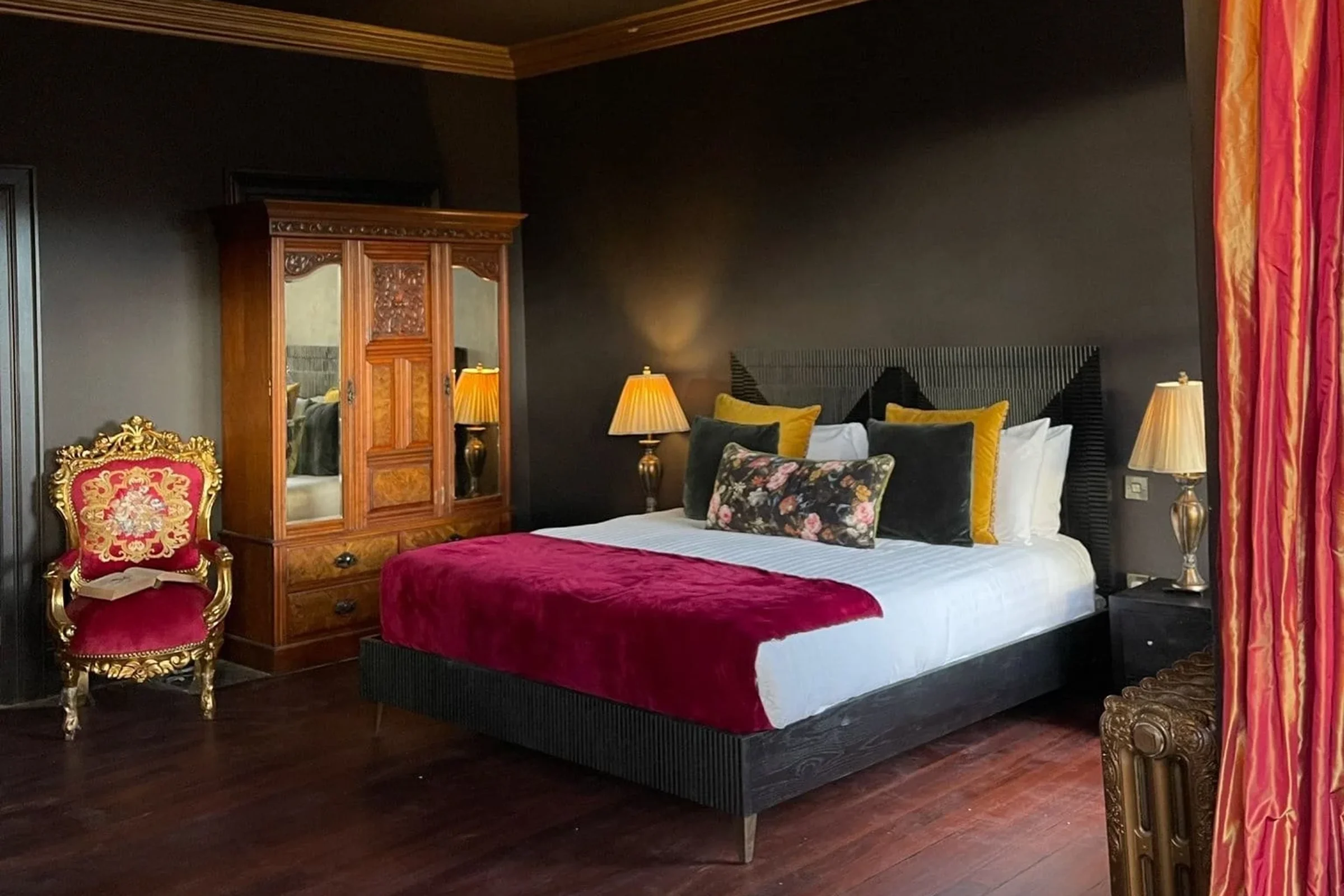 A bedroom with black walls, a black bed frame, and a white bed with multiple pillows, including yellow, dark green, and floral patterns. Two bedside lamps with yellow shades, an ornate red and gold chair, a wooden wardrobe with mirrored doors, and re