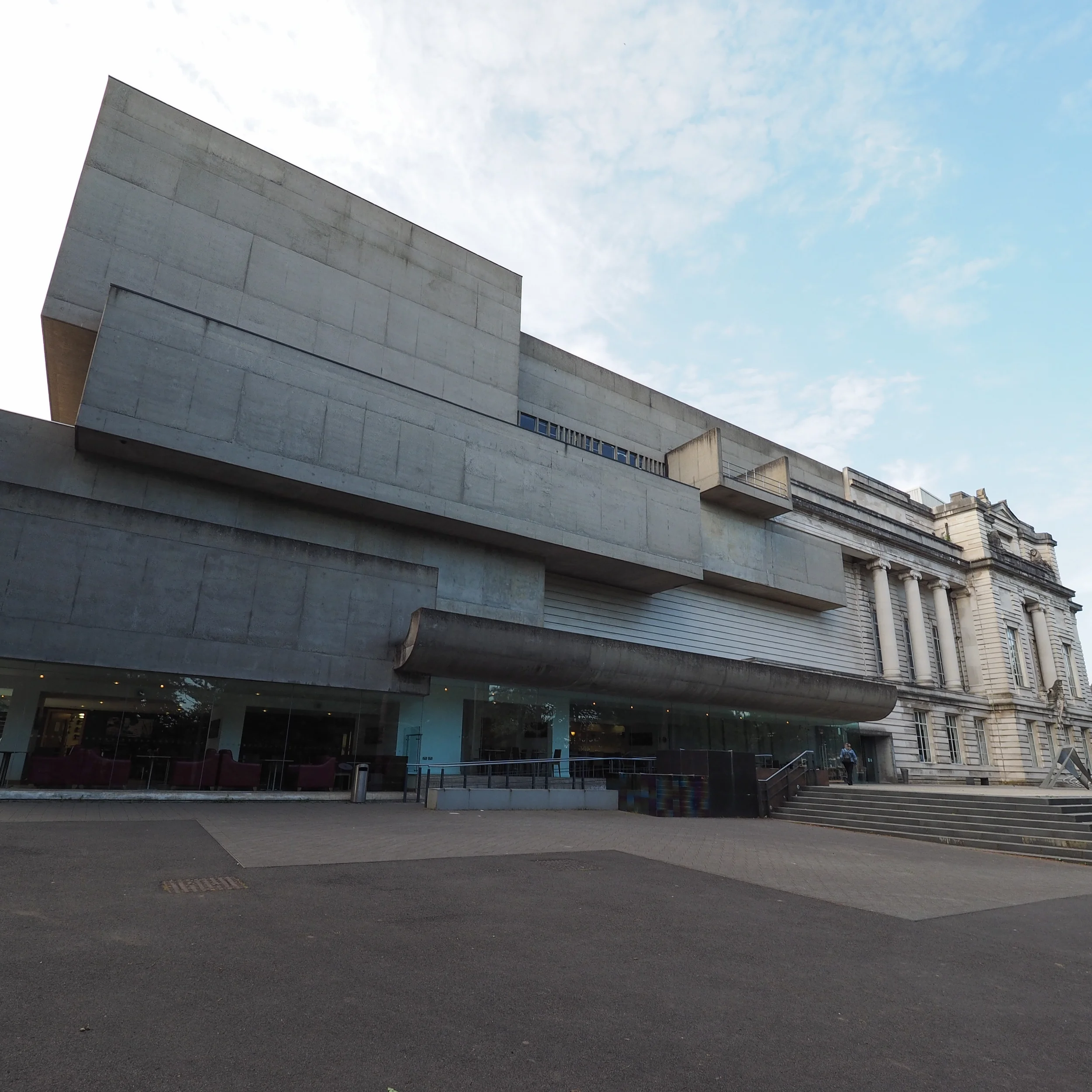 Modern concrete building with large overhangs, steps, and glass windows, adjacent to a historic stone building, against a partly cloudy sky.