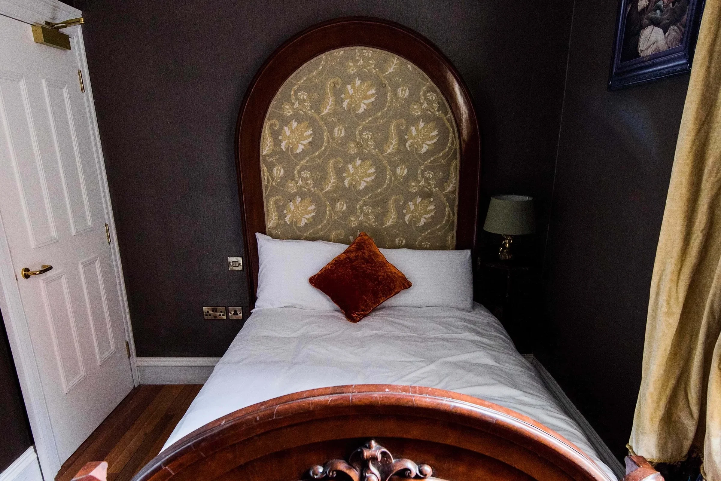 A hotel room with a neatly made bed featuring white sheets, a white pillow, and a brown decorative pillow. The bed has a large, curved headboard with an ornate, patterned fabric inset and a polished wooden frame. To the right of the bed, there is a s