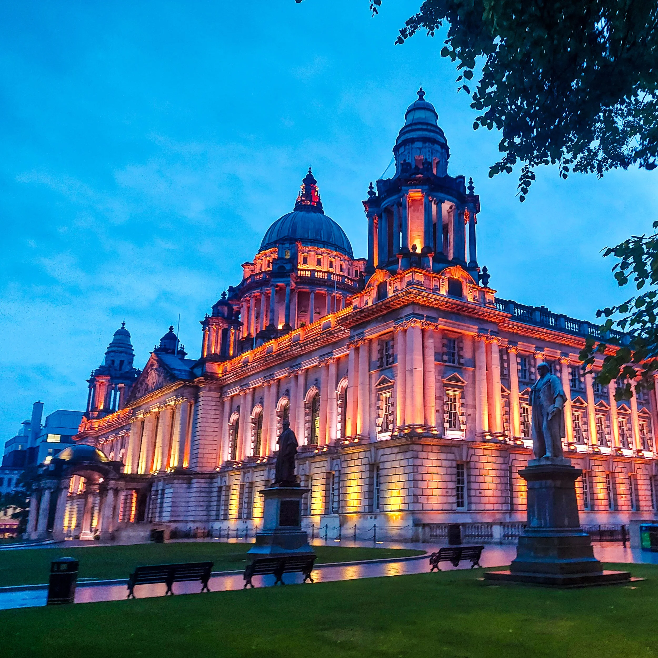 The image shows an illuminated historic building with a large dome and towers, set against a darkening sky, with trees and statues in the foreground.