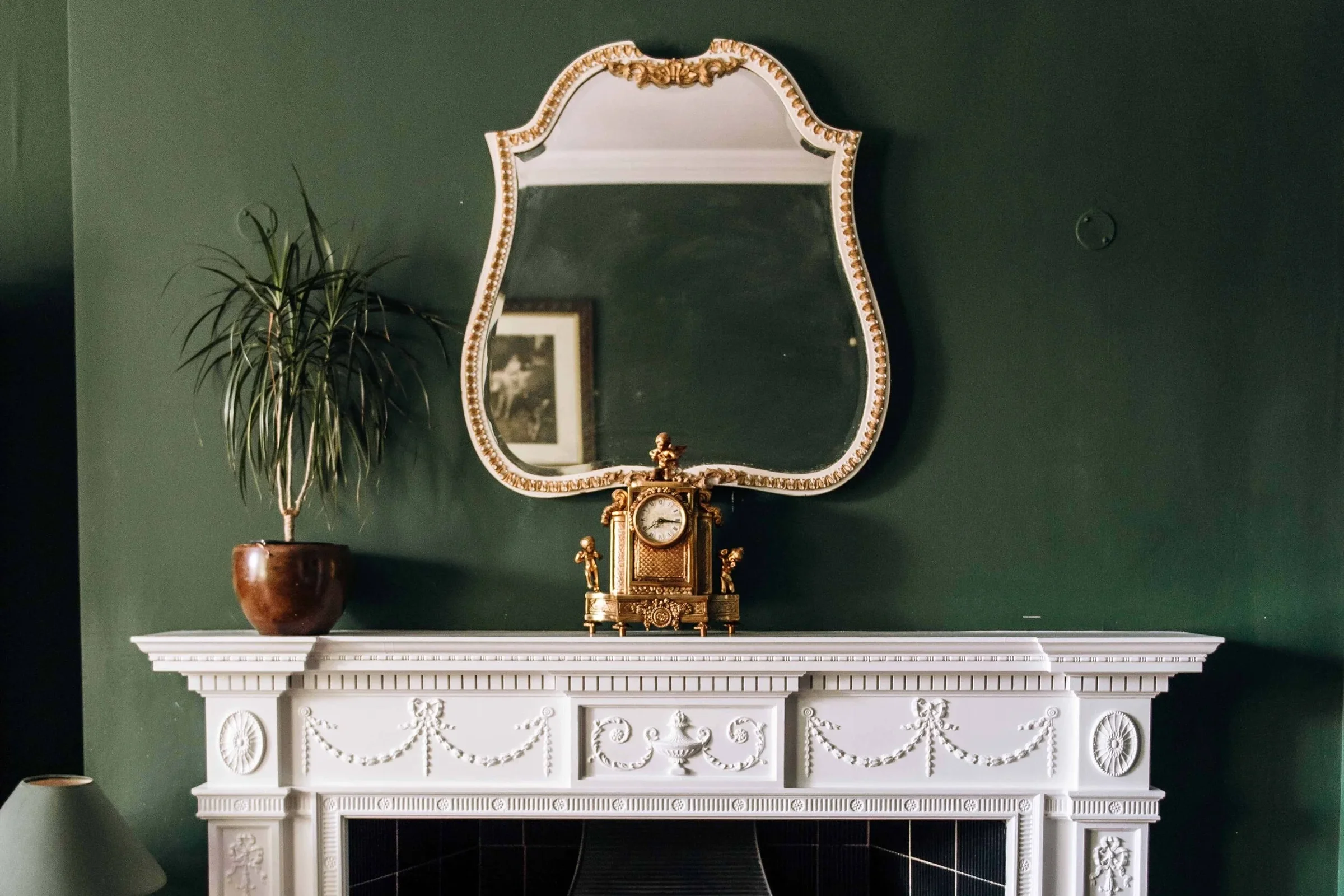 Elegant white fireplace mantel with decorative molding, a vintage gold clock, a large green potted plant on the left, a mirror with a decorative frame above, and a dark green wall background.
