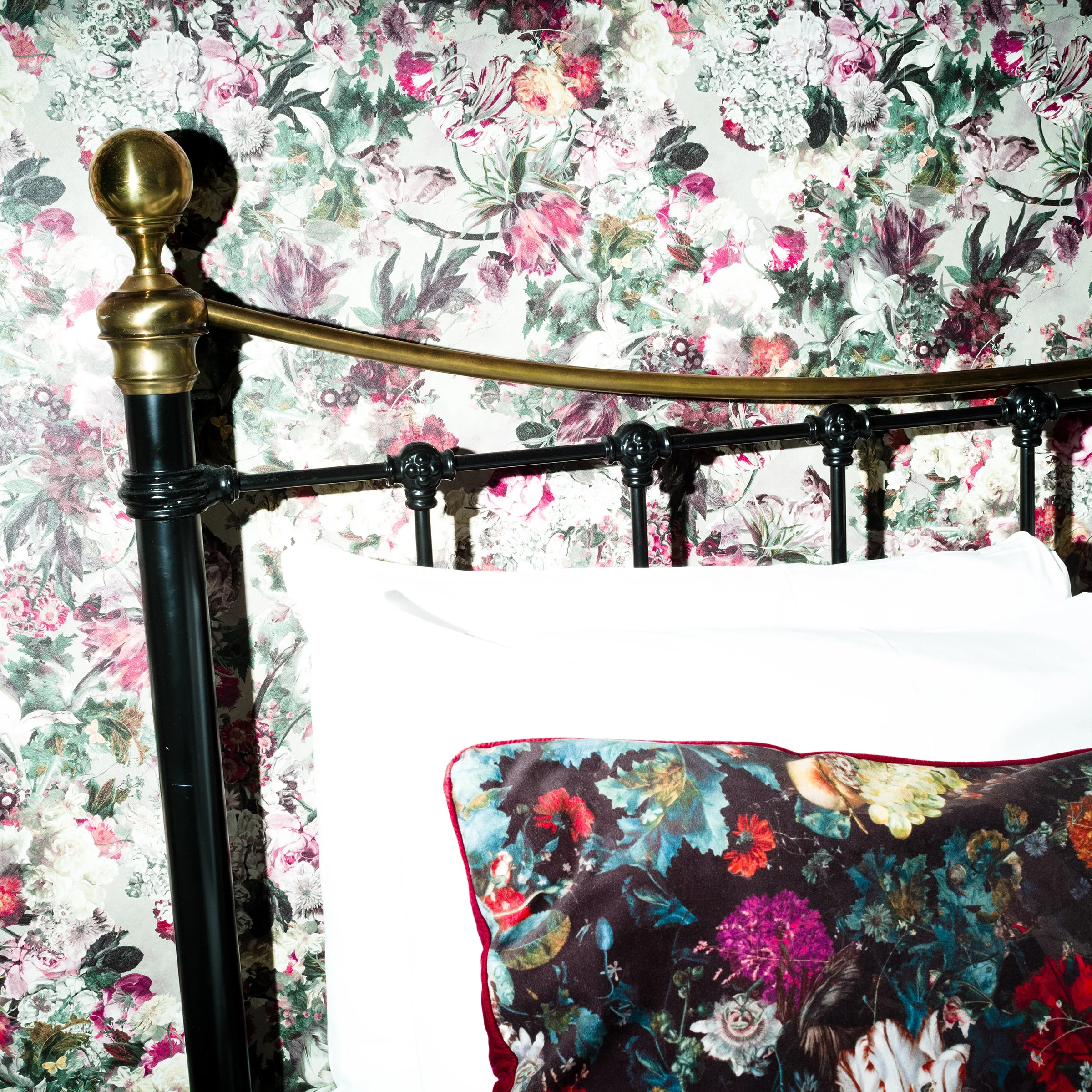Part of a bedroom with a floral wallpaper, a black metal bed frame with brass details, white bedding, and a floral pillow that matches the wallpaper.
