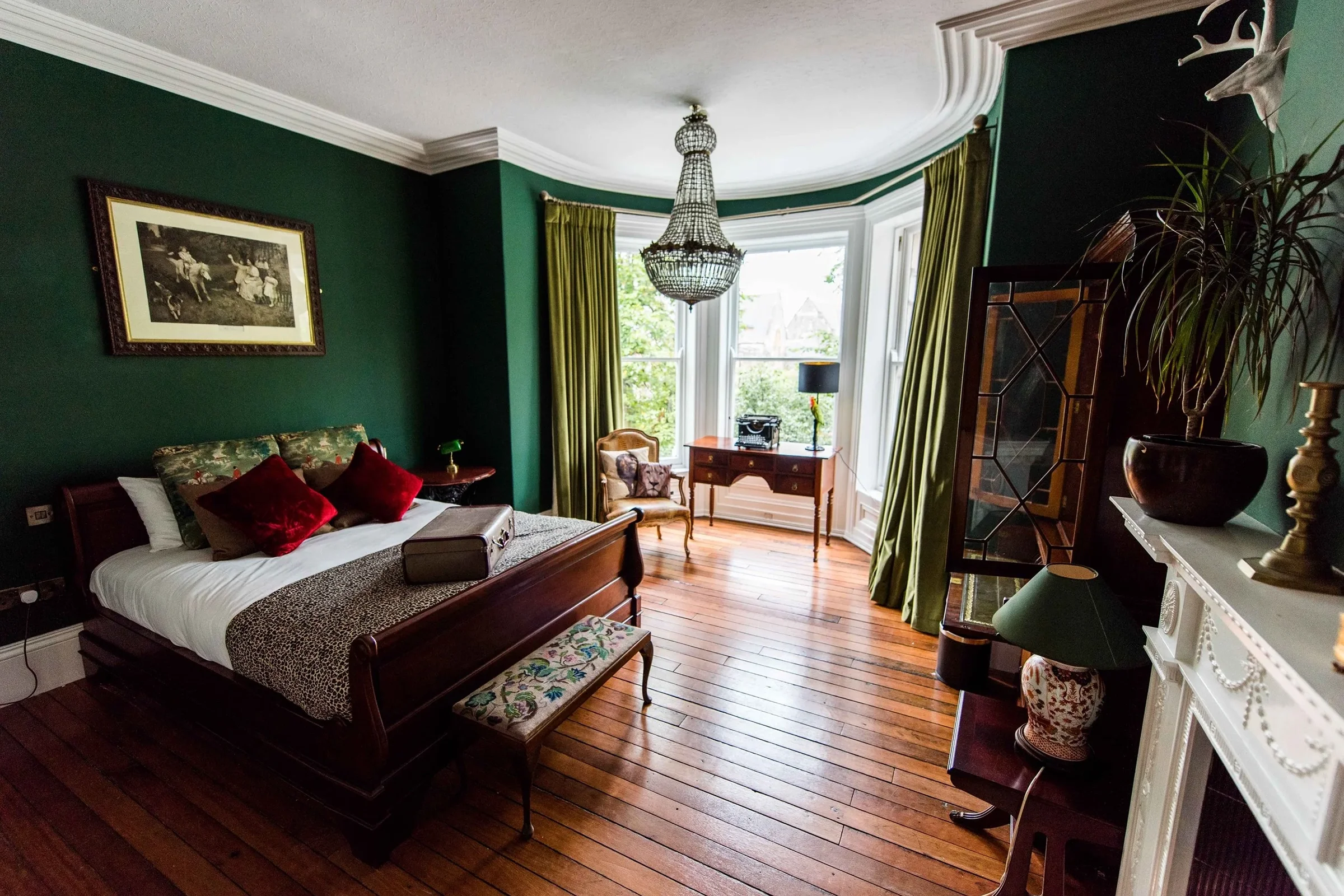 A vintage bedroom with green walls, a large bed with animal print and velvet pillows, wooden floor, and a bay window with green curtains. Decor includes a chandelier, painting, and antique furniture.