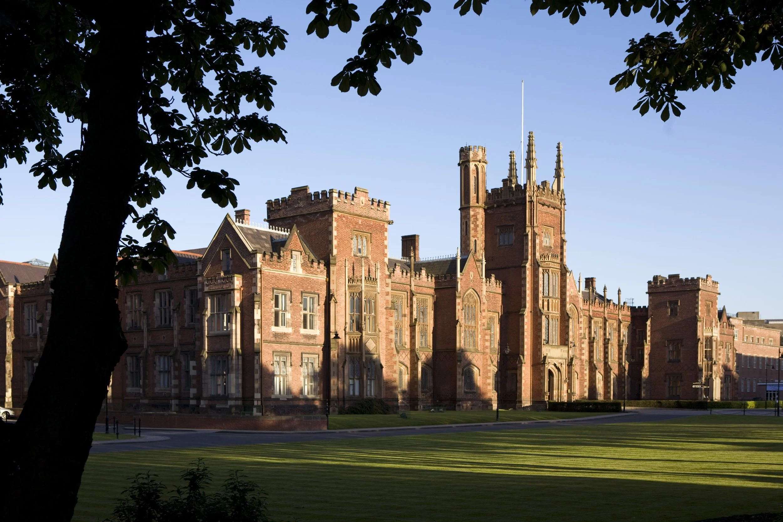 A large historic castle with red brick walls, towers, and detailed architectural features, surrounded by a green lawn and framed by trees.