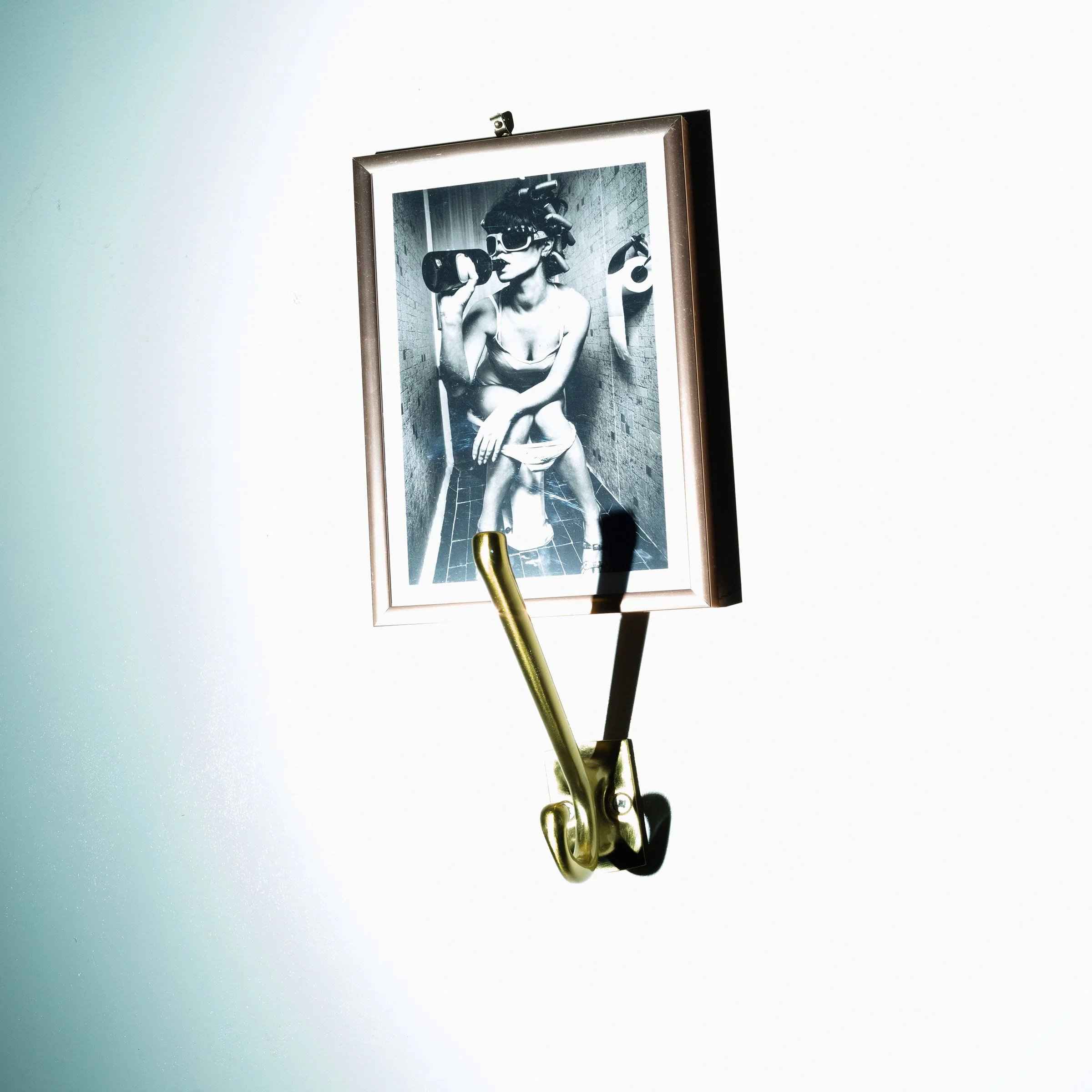 Photograph of a framed black and white artwork hung on a white wall, depicting a woman in a bathroom wearing goggles, holding a dumbbell, and sitting on a toilet, with a roll of toilet paper nearby and a toilet paper holder.