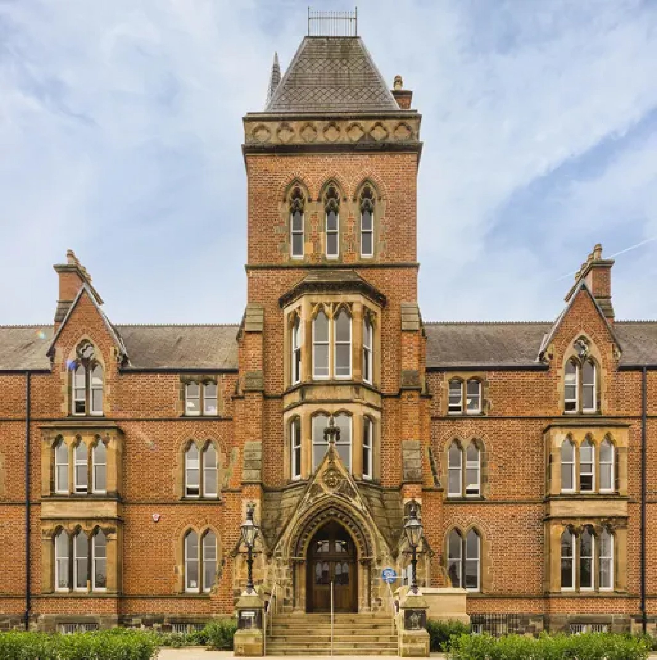 Historic brick building with a tower, arched and rectangular windows, and ornate stone entrance with stairs and lamp posts.