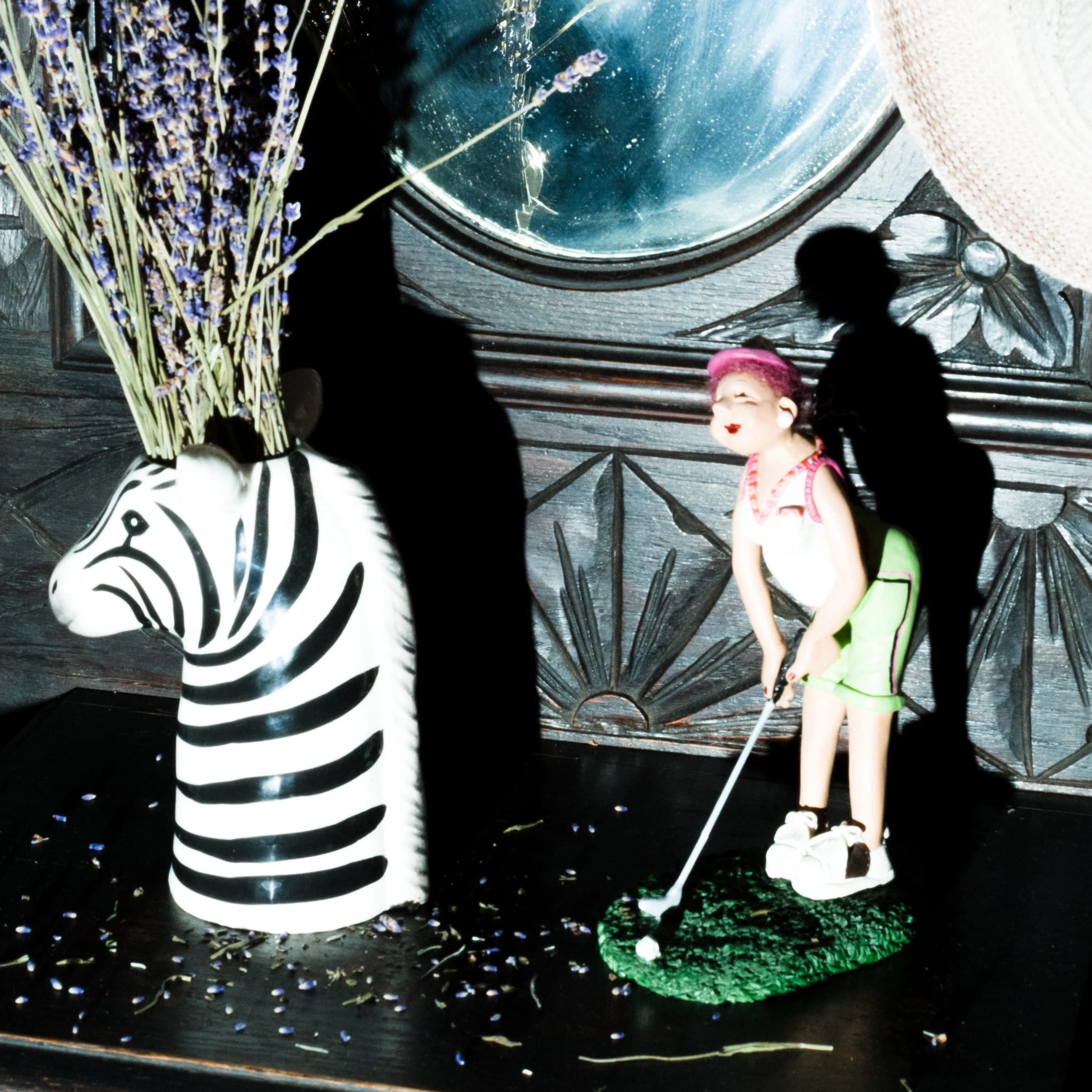 Decorative scene with a zebra-shaped vase holding dried lavender, a figurine of a woman in golfer attire, and shadows cast by objects on a dark surface with a textured wooden background and a round mirror.