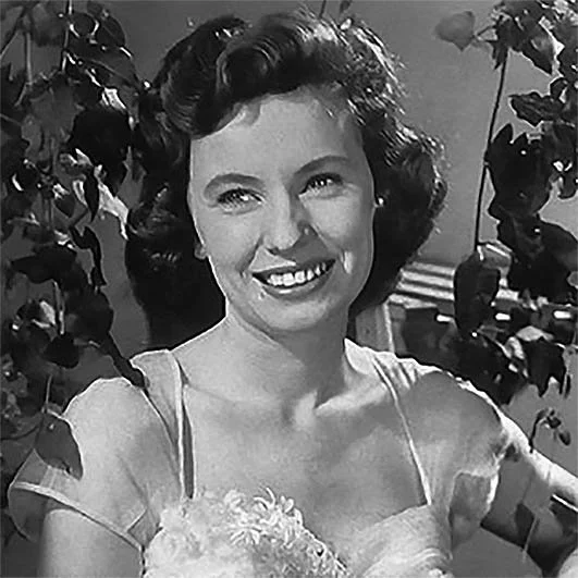 A woman with dark, curly hair smiling, wearing a dress with floral embellishment, standing outdoors with plants in the background.