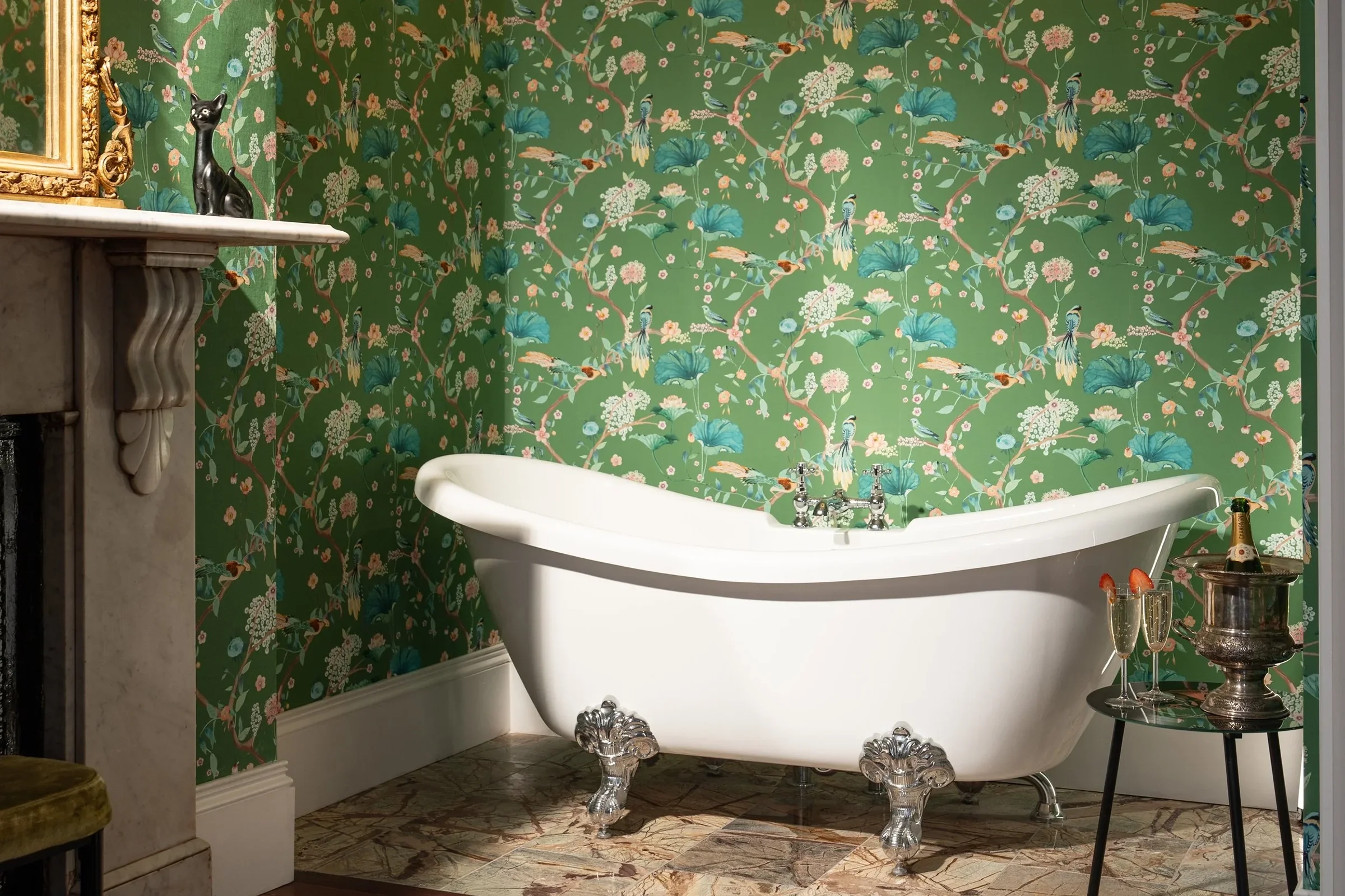 A vintage-style bathroom with a white clawfoot bathtub, floral wallpaper, a small table with champagne and glasses, and a fireplace with decorative items.