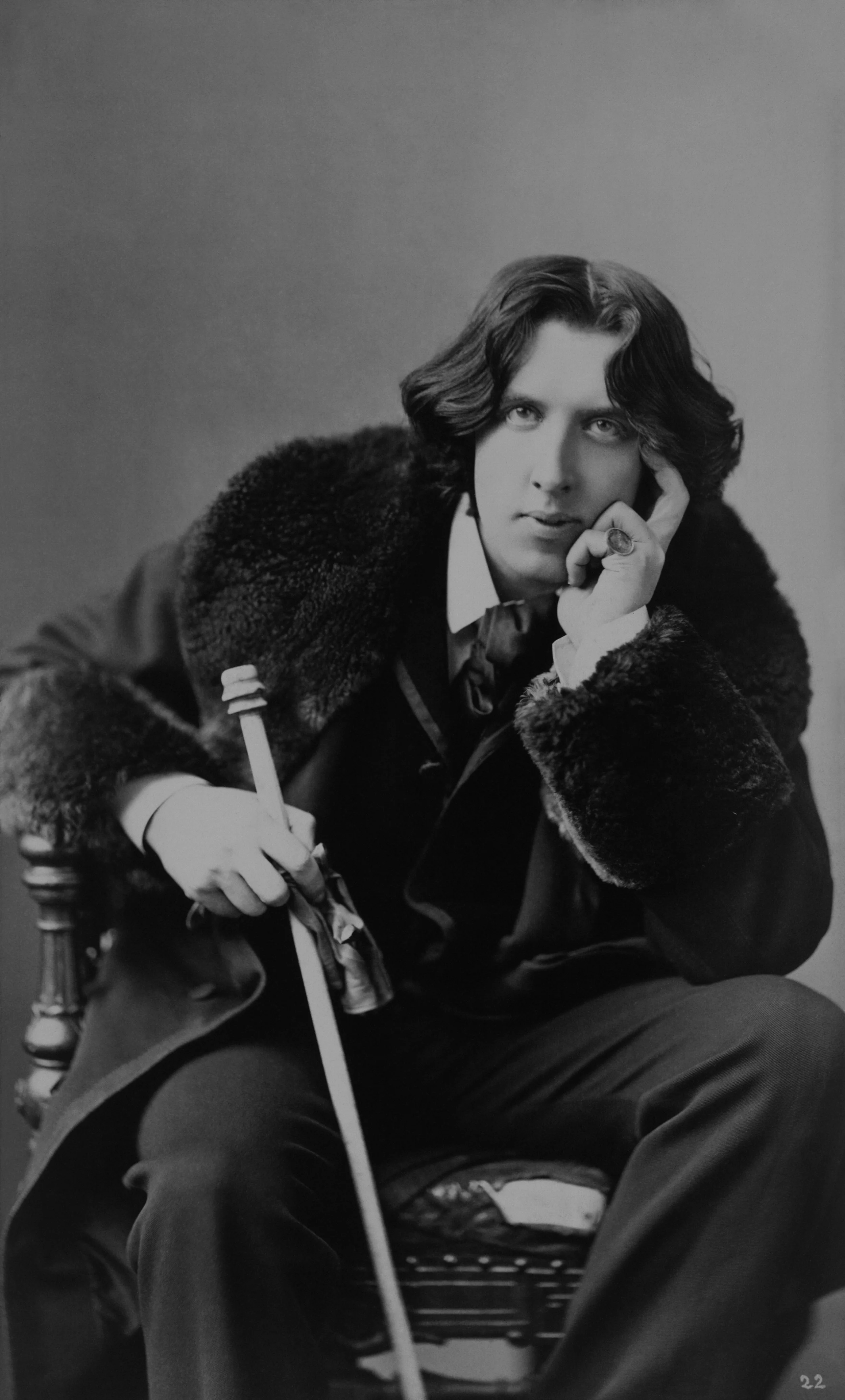 Black and white portrait of a man with wavy hair, wearing a luxurious coat with a fur collar, sitting casually with a cane, in a thoughtful pose.