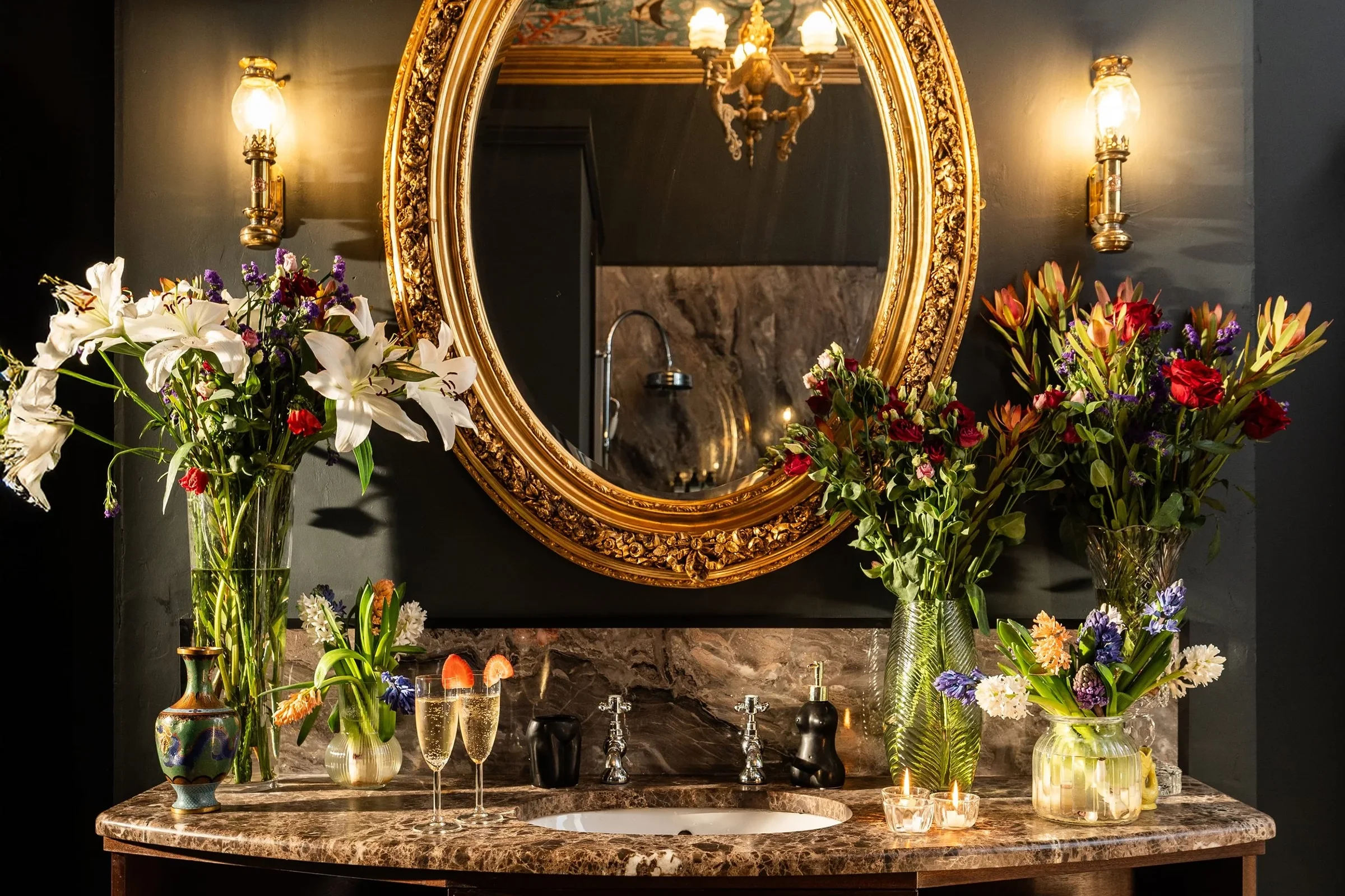 A luxurious bathroom vanity with an ornate gold-framed mirror, dark walls, and a marble countertop. The countertop holds various flower arrangements in vases, candles, and glassware. Two wall sconces with glass shades provide warm lighting.