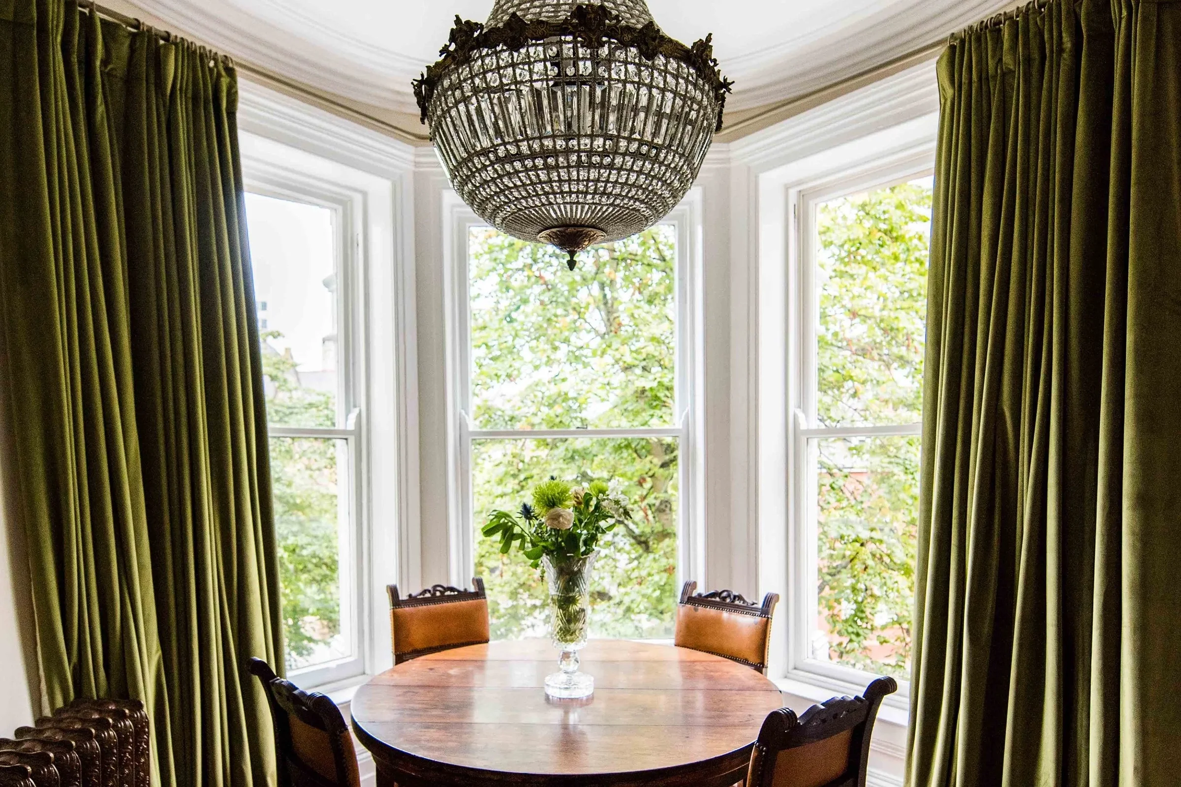 Interior of a dining room with a round wooden table, four chairs, a floral centerpiece, large bay windows with green curtains, and a chandelier hanging from the ceiling.