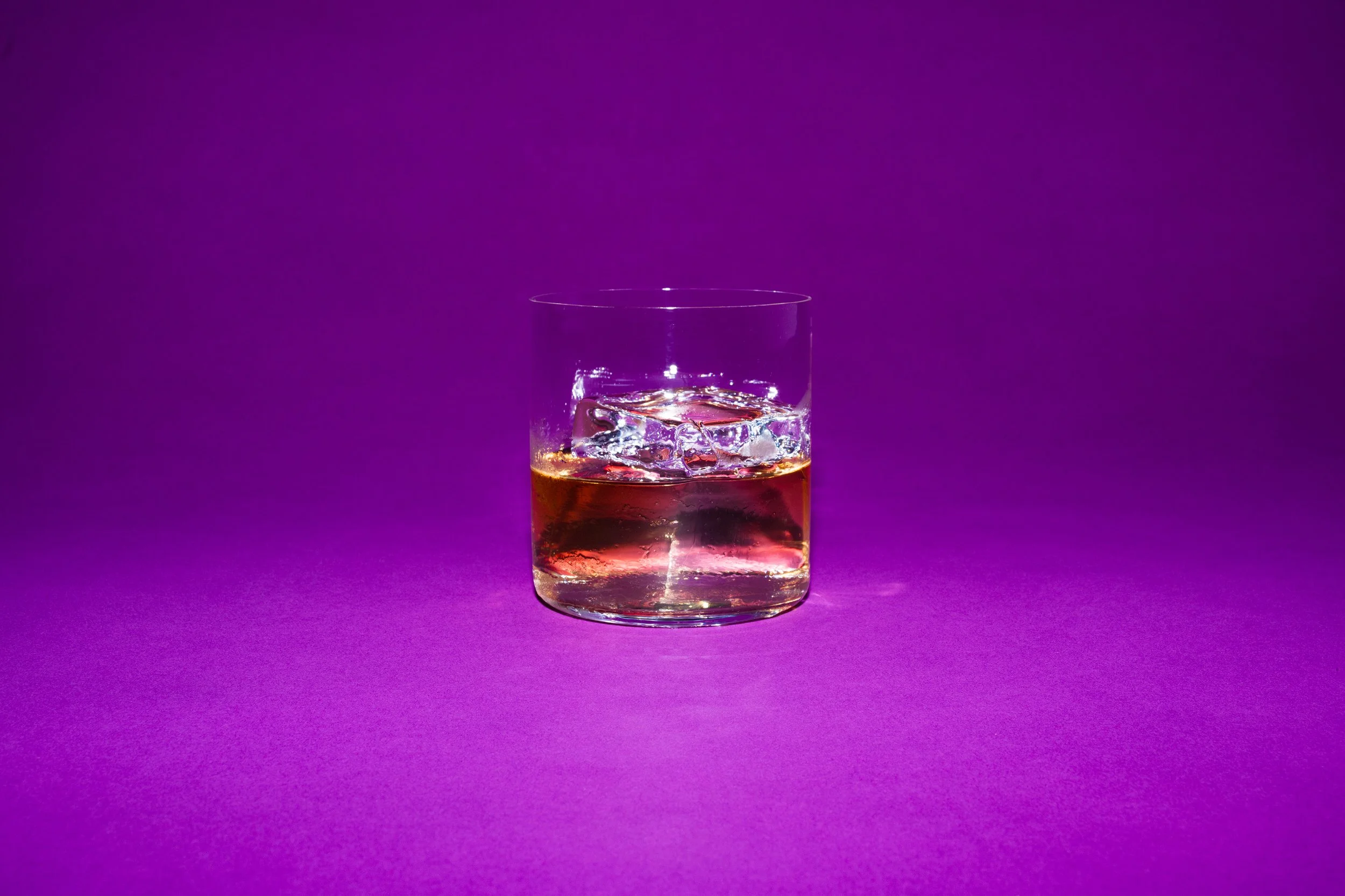 A glass of whiskey with ice cubes against a purple background.