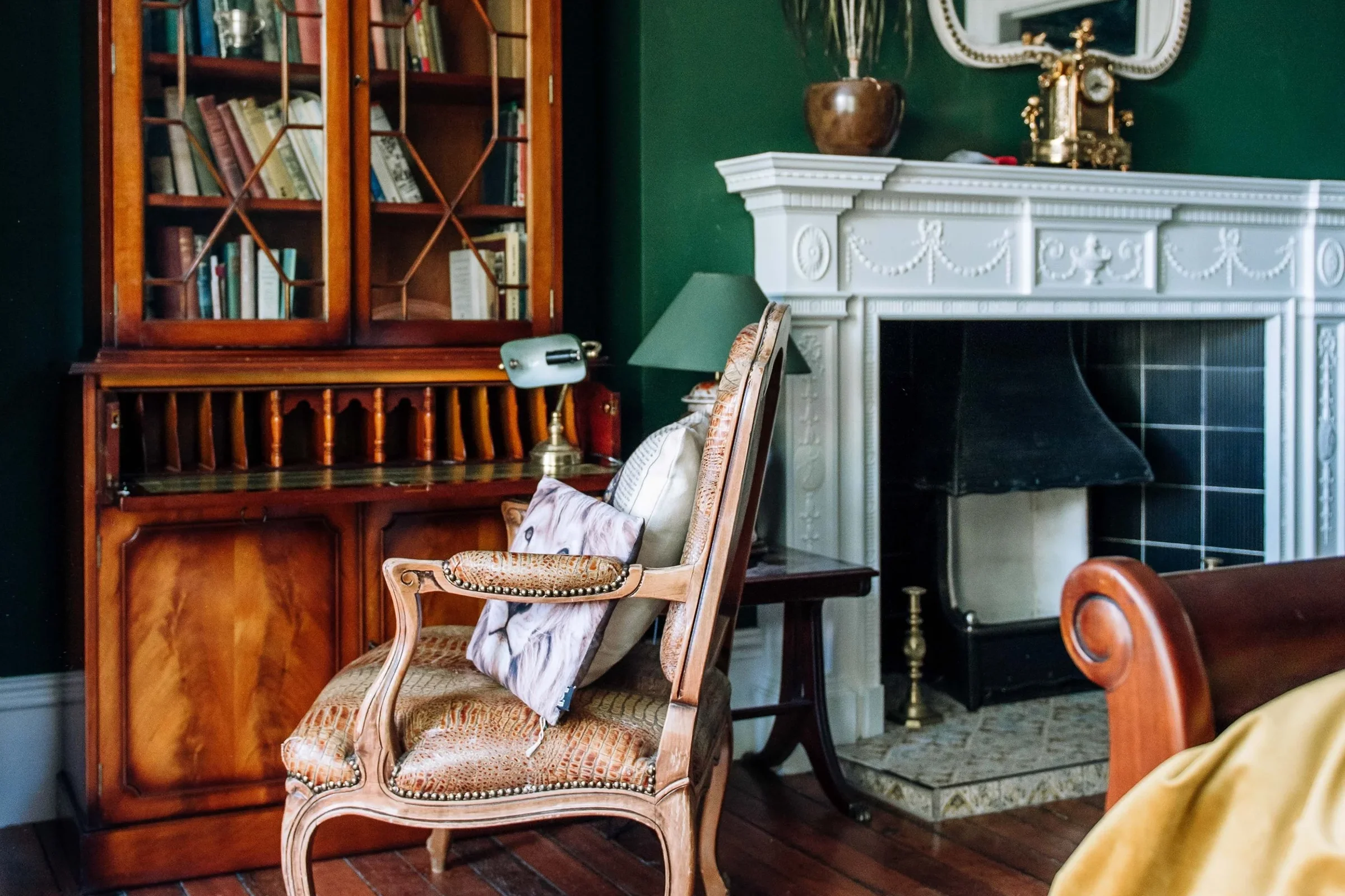 A cozy living room corner with a wooden bookshelf, a cushioned armchair with pillows, a lamp, and a fireplace with decorative trim.