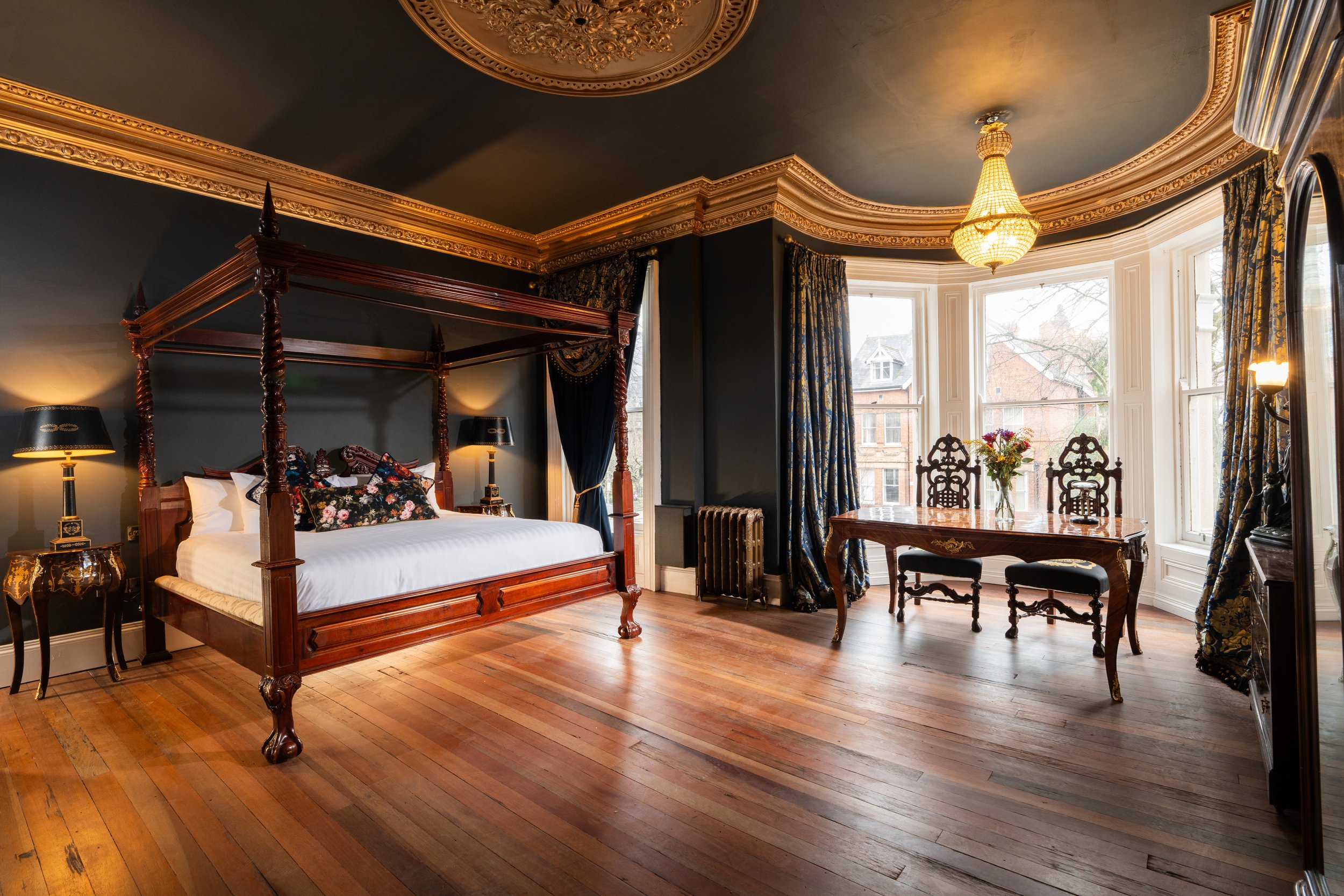 Elegant bedroom with a four-poster bed, hardwood floors, and a bay window with floral curtains, featuring a small table with a flower vase.