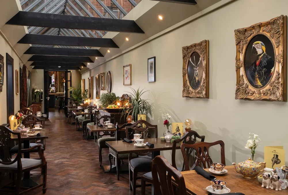 Interior of a cozy, elegant restaurant with wooden tables and chairs, framed portraits on the wall, and decorative plants.