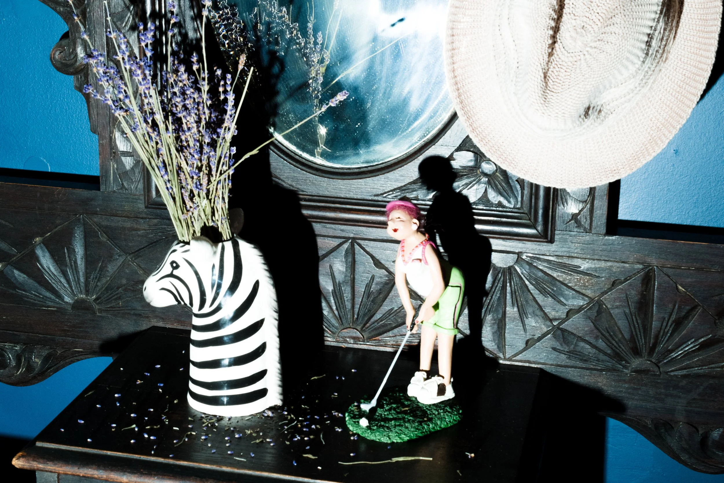 Decorative shelf with a zebra-shaped vase holding purple lavender flowers, a small figurine of a girl in golf attire with pink hair, wearing white shoes, standing on a green base, and a large woven hat hanging above. Behind the shelf is an ornate wooden mirror frame and a blue wall.