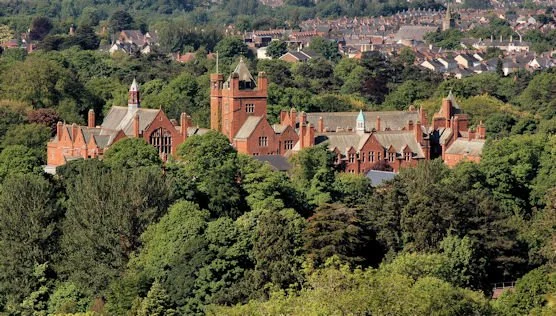 A large historic red brick building with towers surrounded by dense green trees, with a residential neighborhood in the background.