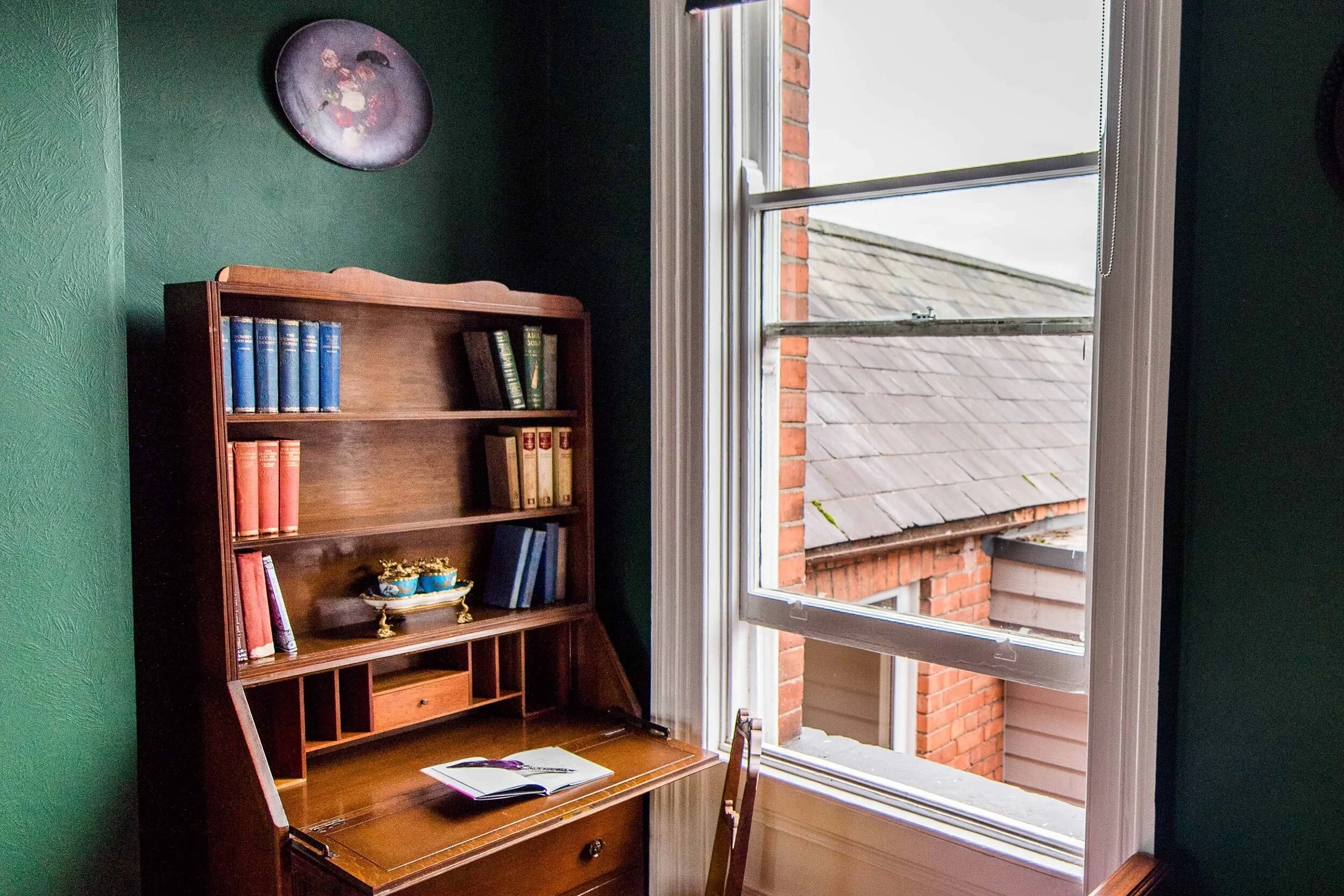 A wooden secretary desk with a slanted writing surface, open to reveal small compartments, situated next to an open window with a view of brick rooftops.