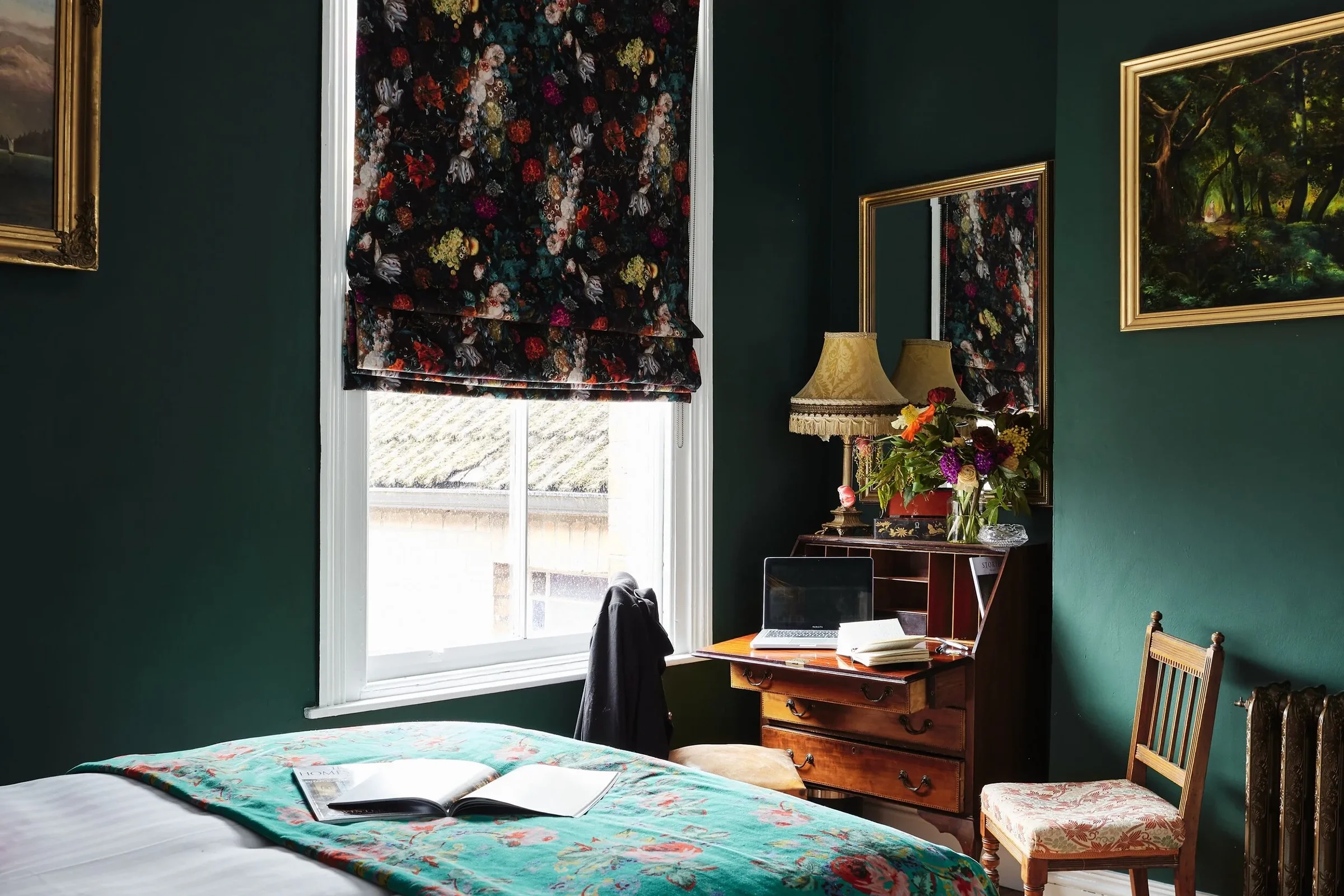 A cozy bedroom with dark green walls, a window with floral window shades, a bed with a colorful blanket, a wooden desk with a laptop and books, a vintage chair with floral upholstery, and paintings on the walls.