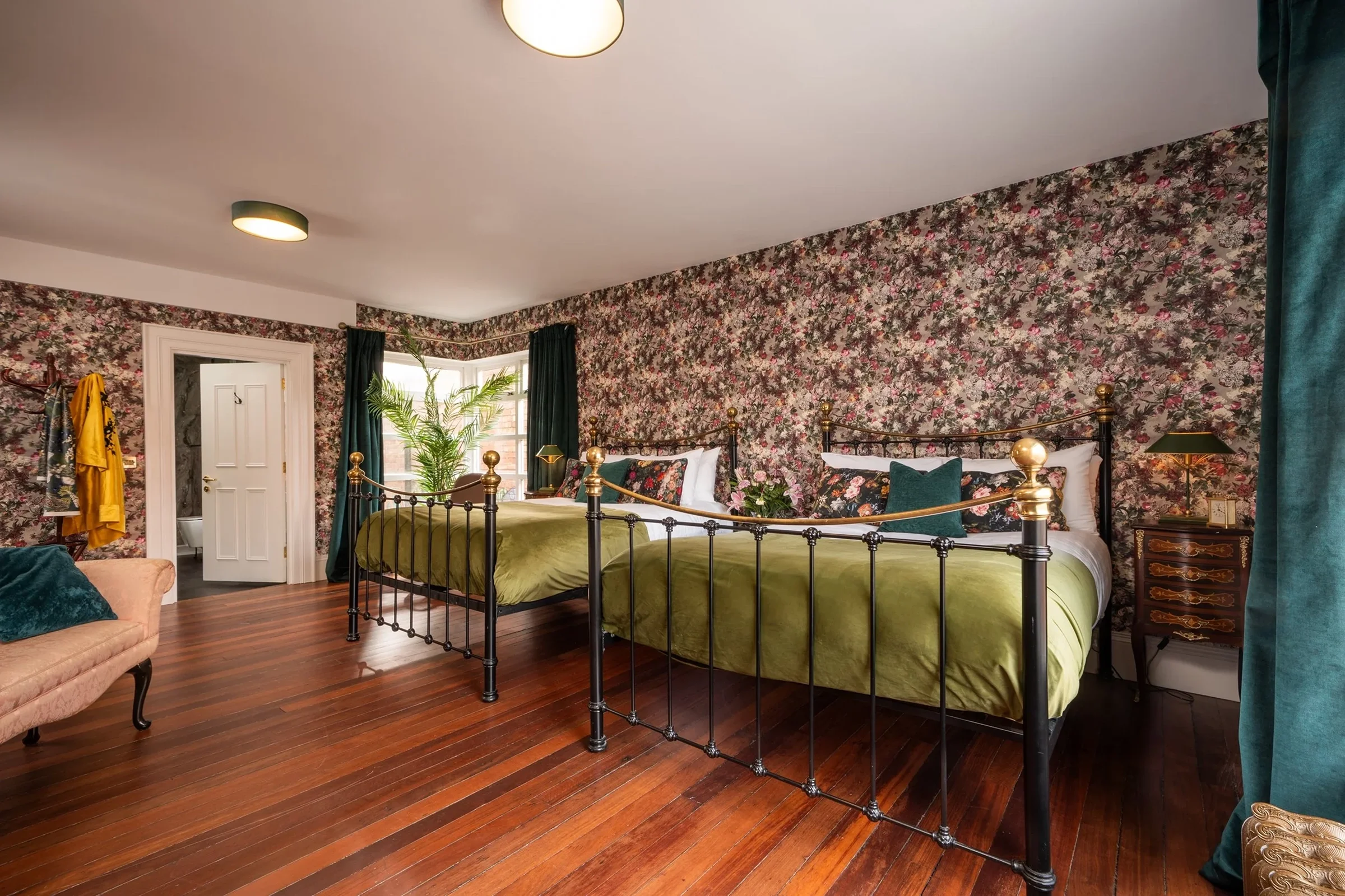 A bedroom with floral wallpaper, two green beds with black metal frames, a brown nightstand, and a pink armchair next to a teal curtain. The room has wooden floors and a large window with dark green curtains.