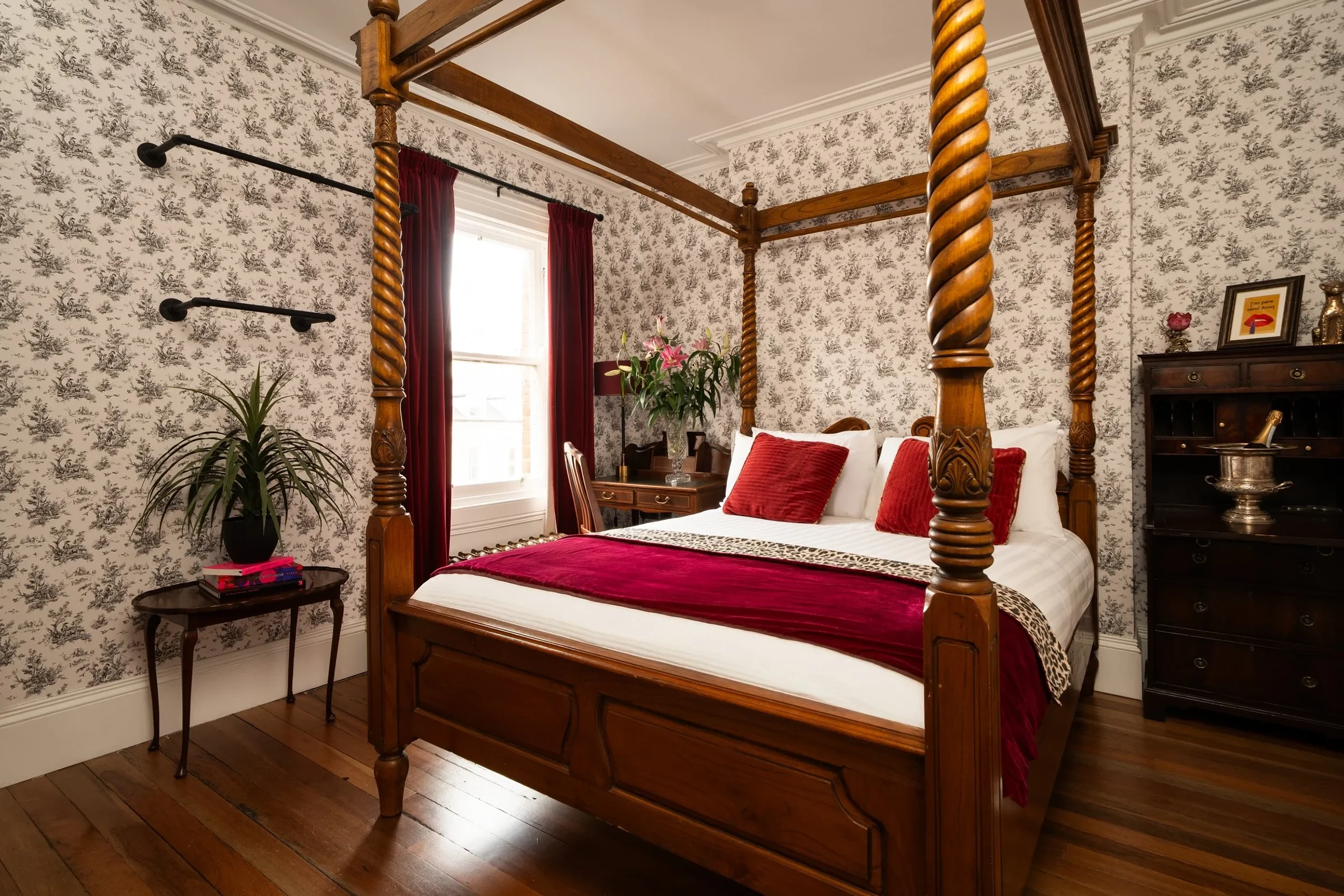 A bedroom with a wooden four-poster bed, floral wallpaper, and red accents, including pillows, curtains, and a blanket. There is a side table with books and a plant, and a dresser with decorative items.