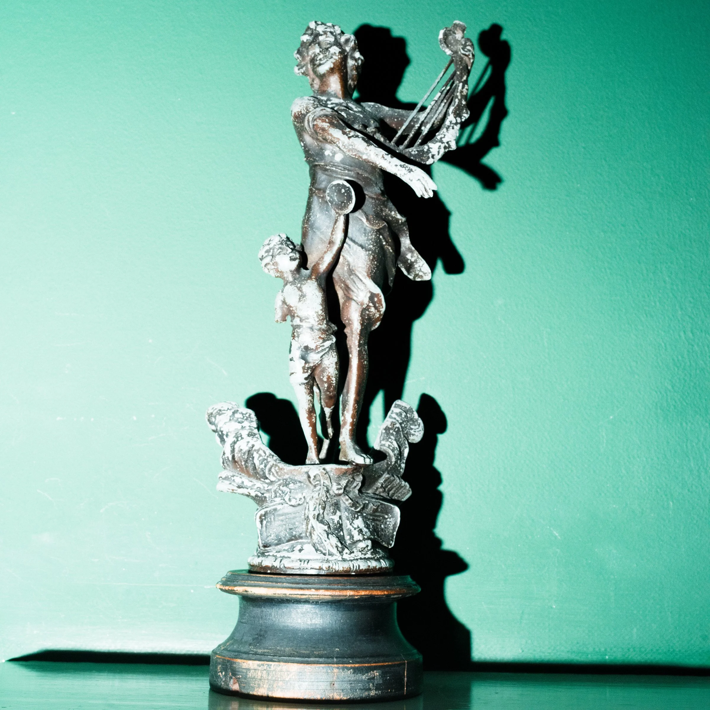 A silver sculpture of a woman holding a bow and arrow, with a child holding her hand at her side, standing on a circular base against a green background with a shadow.