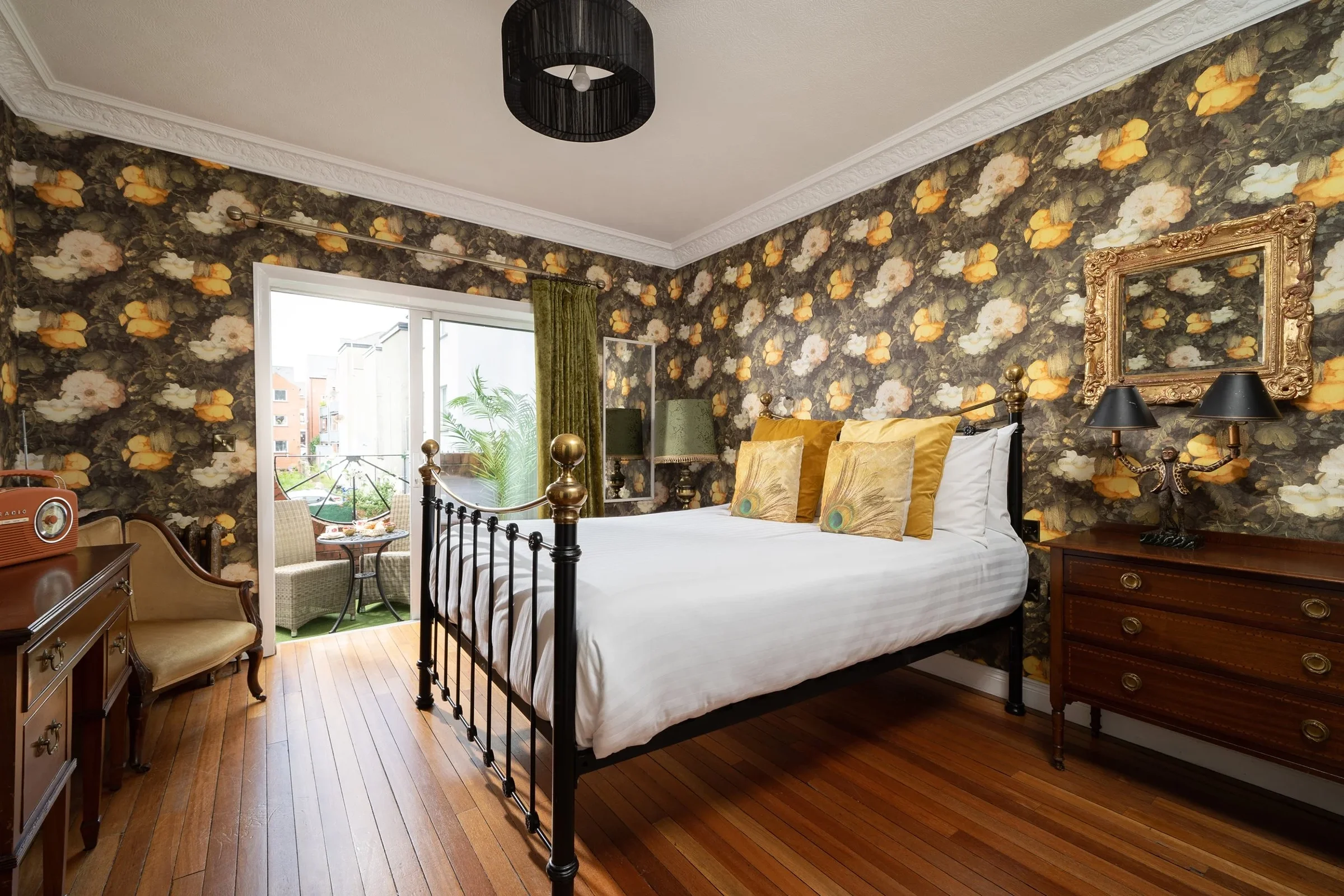 A bedroom with floral wallpaper, a bed with peacock feather pillows, and an outdoor balcony with seating