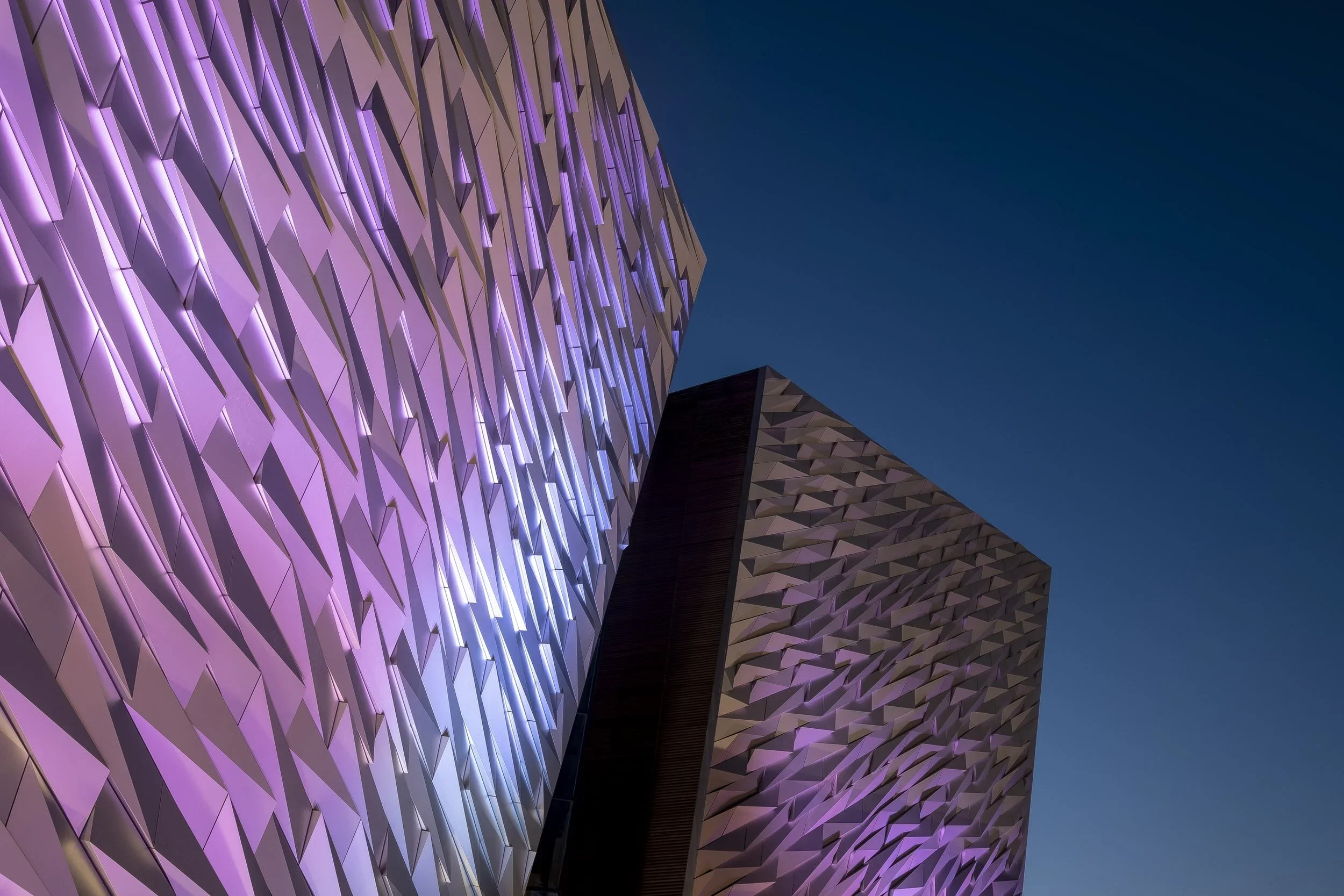 Nighttime view of two modern buildings with geometric, textured facades illuminated by purple lights against a dark blue sky.