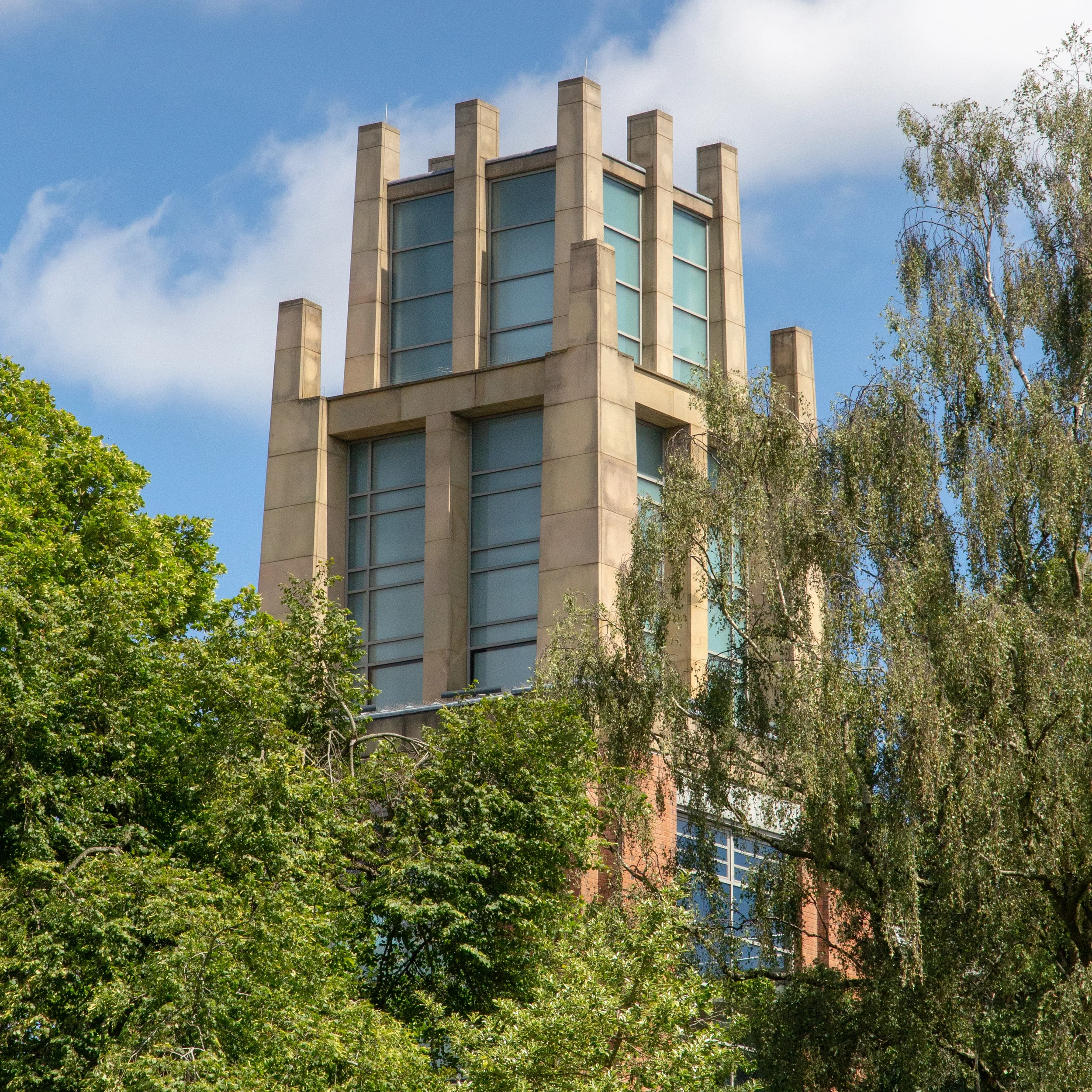 A tall, modern building with a beige facade and large glass windows, partially obscured by green trees, with a blue sky and some clouds in the background.