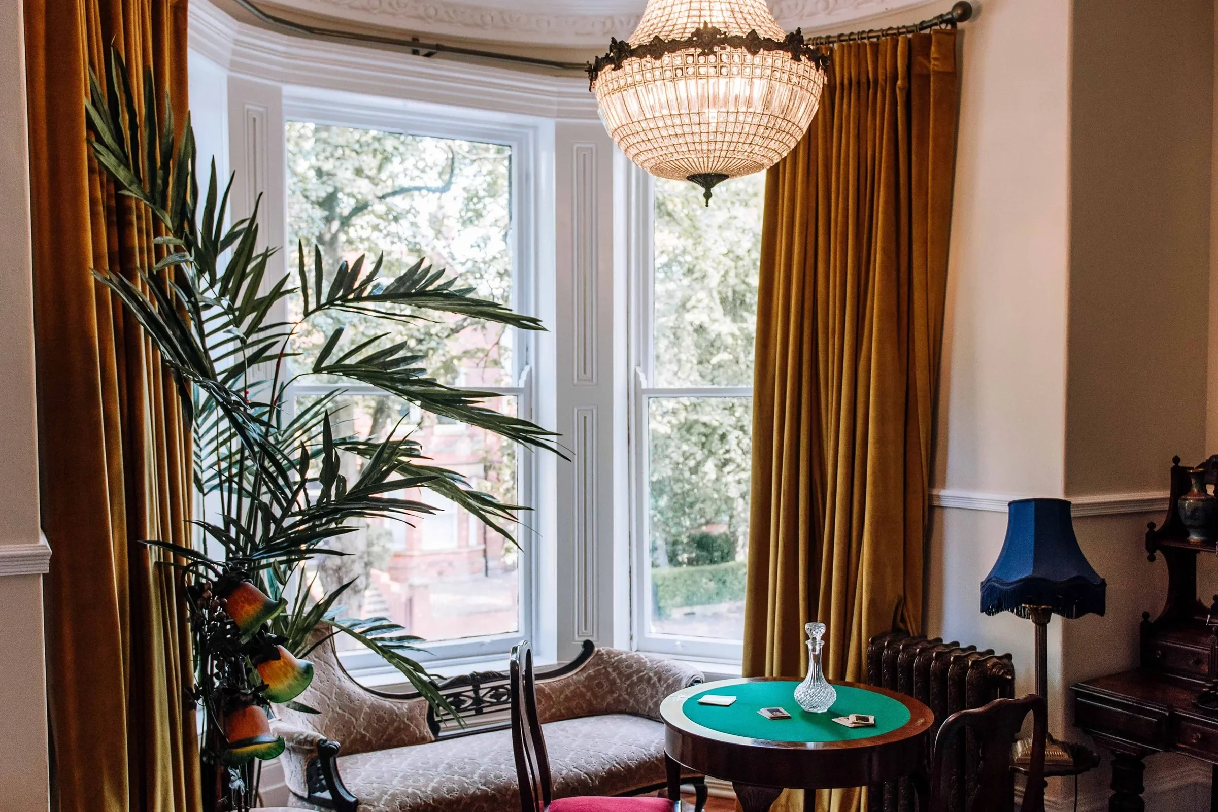 Vintage living room with large bay window, gold curtains, a chandelier, a small round table with playing cards and a glass vase, a blue lamp, a cushioned bench, and a tall green plant.