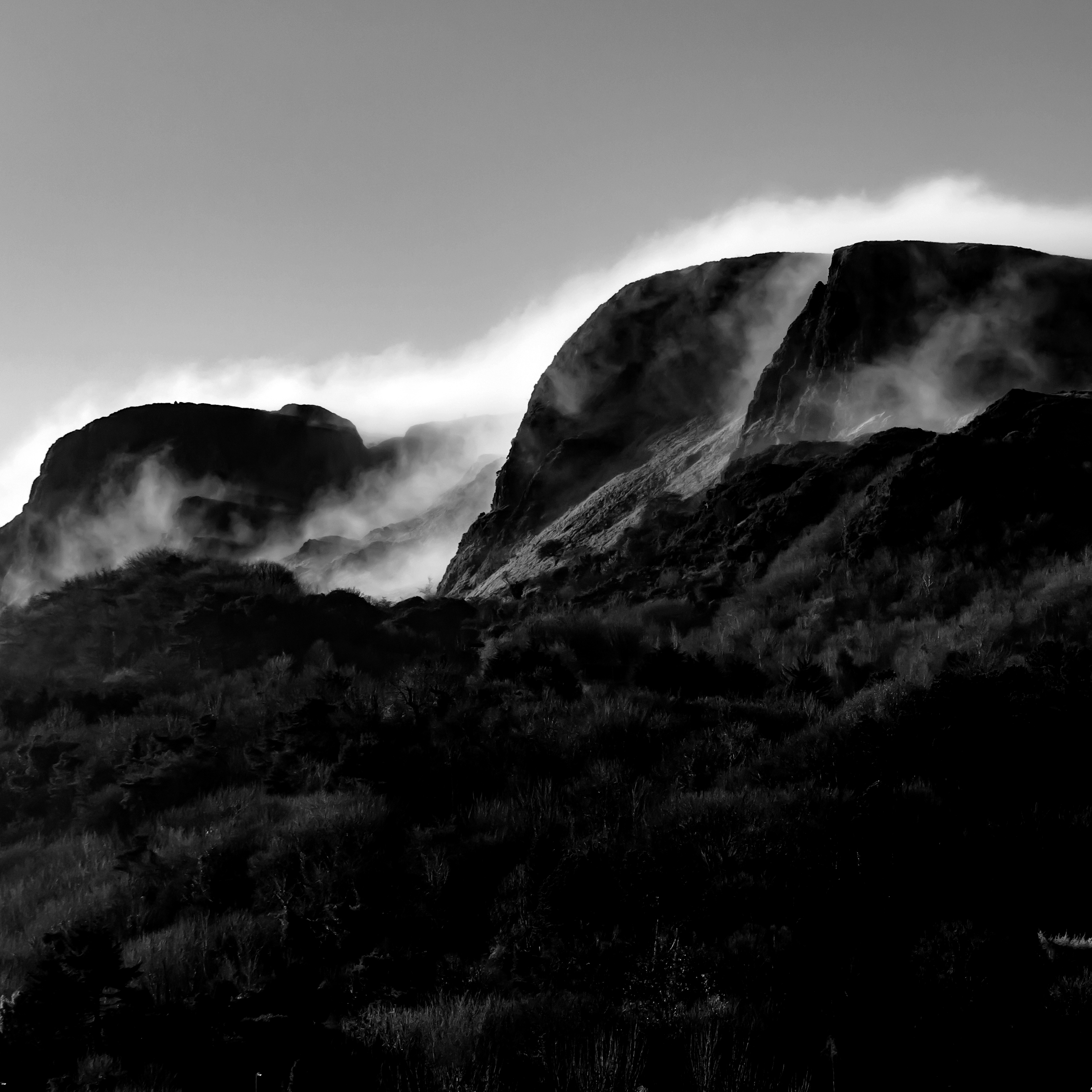 Black and white photo of rugged mountains with mist or clouds surrounding them and sparse vegetation in the foreground.