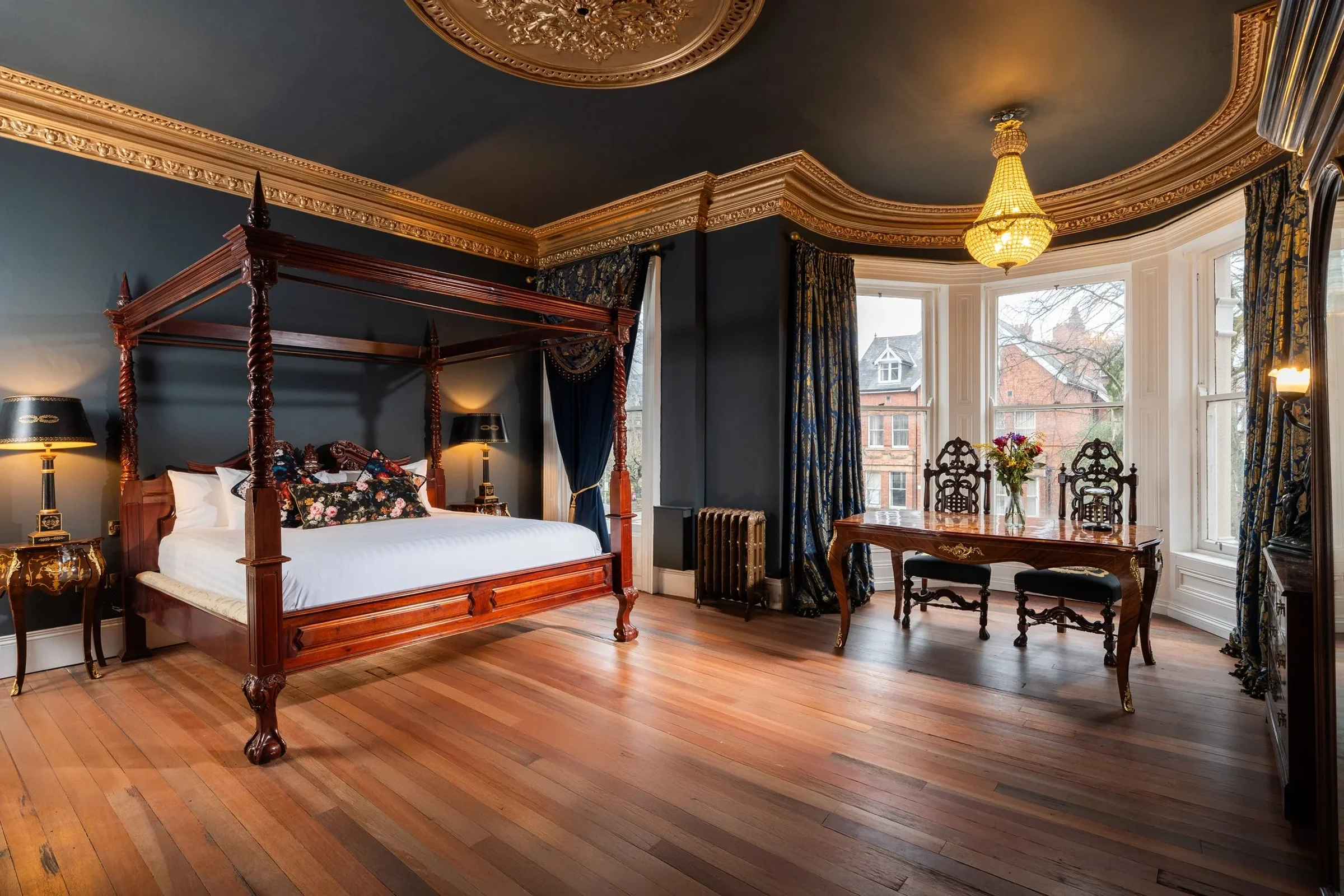 A luxurious bedroom with dark blue walls, a wooden four-poster bed with intricate carvings, matching side tables with lamps, a round wooden table with a floral arrangement, large windows with heavy blue and gold patterned curtains, hardwood flooring,