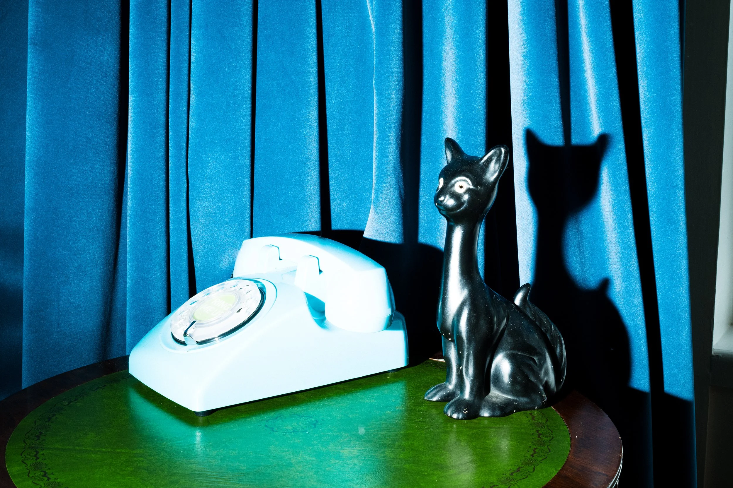 A white rotary telephone and a black ceramic cat figurine on a green round table with blue curtains in the background.