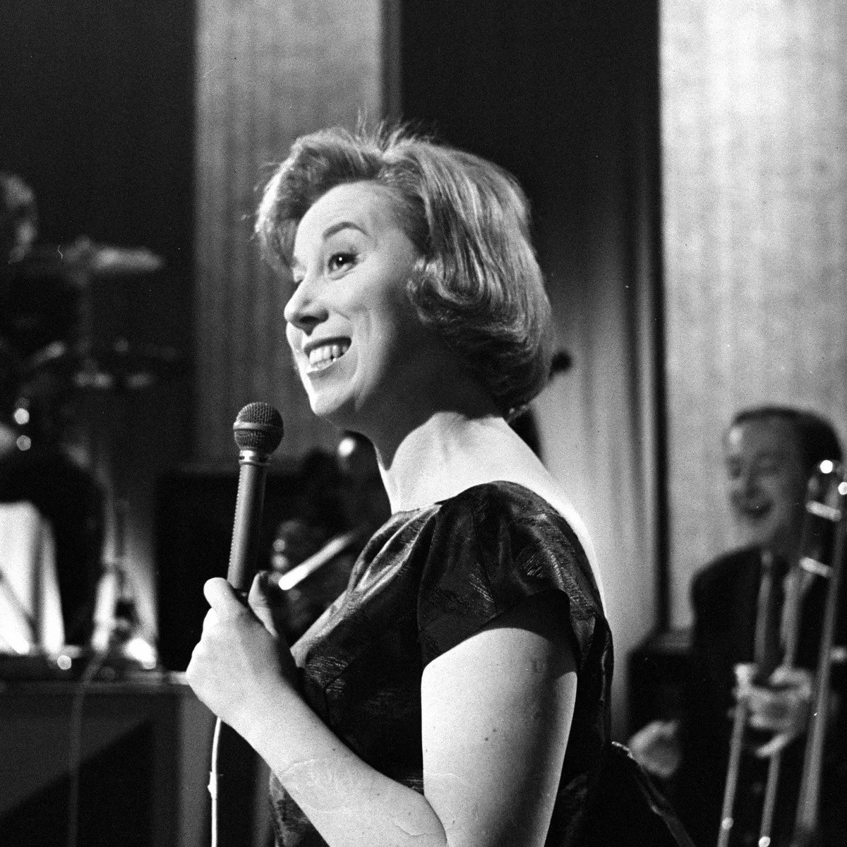 A woman with short hair smiling while holding a microphone, performing on stage, with a band playing in the background.