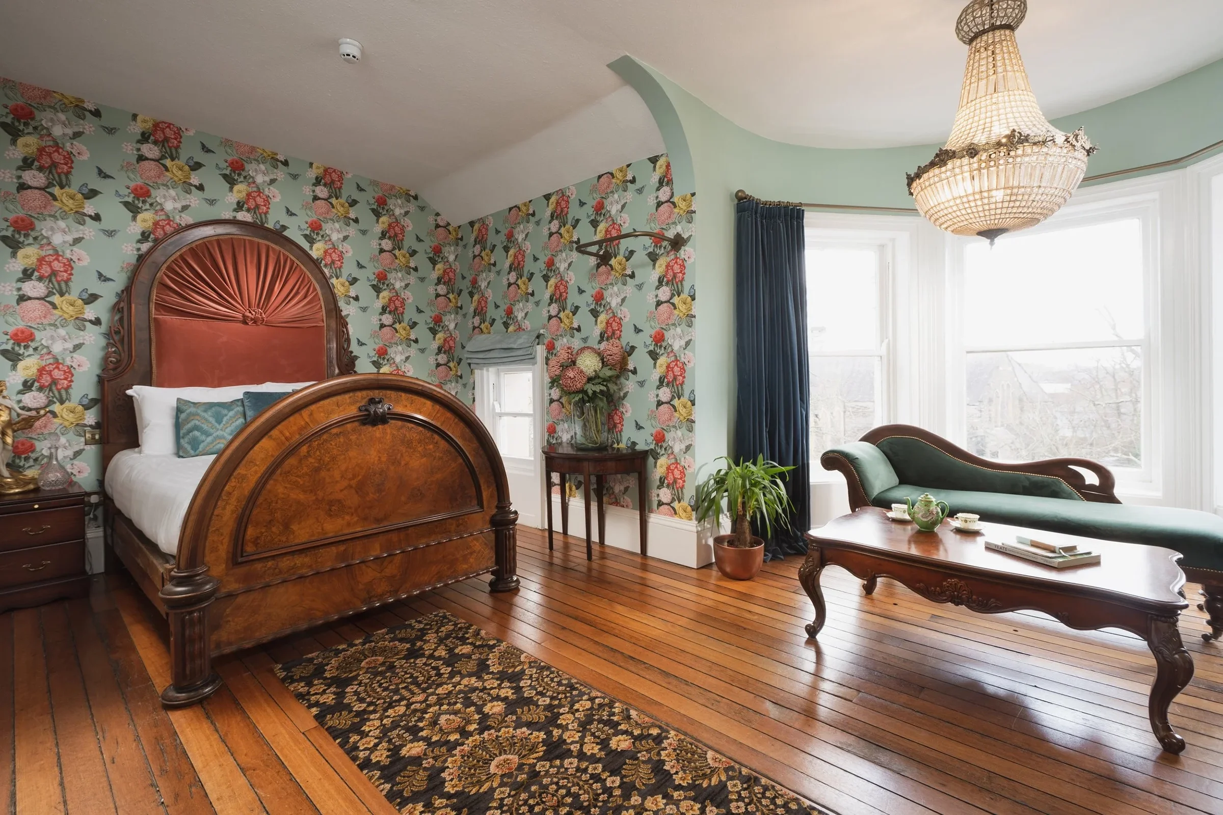 A vintage bedroom with floral wallpaper, a wooden bed with an upholstered headboard, a green chaise lounge, and a chandelier hanging from the ceiling. The room has large windows with blue curtains, wooden floors, and a small table with a flower vase.