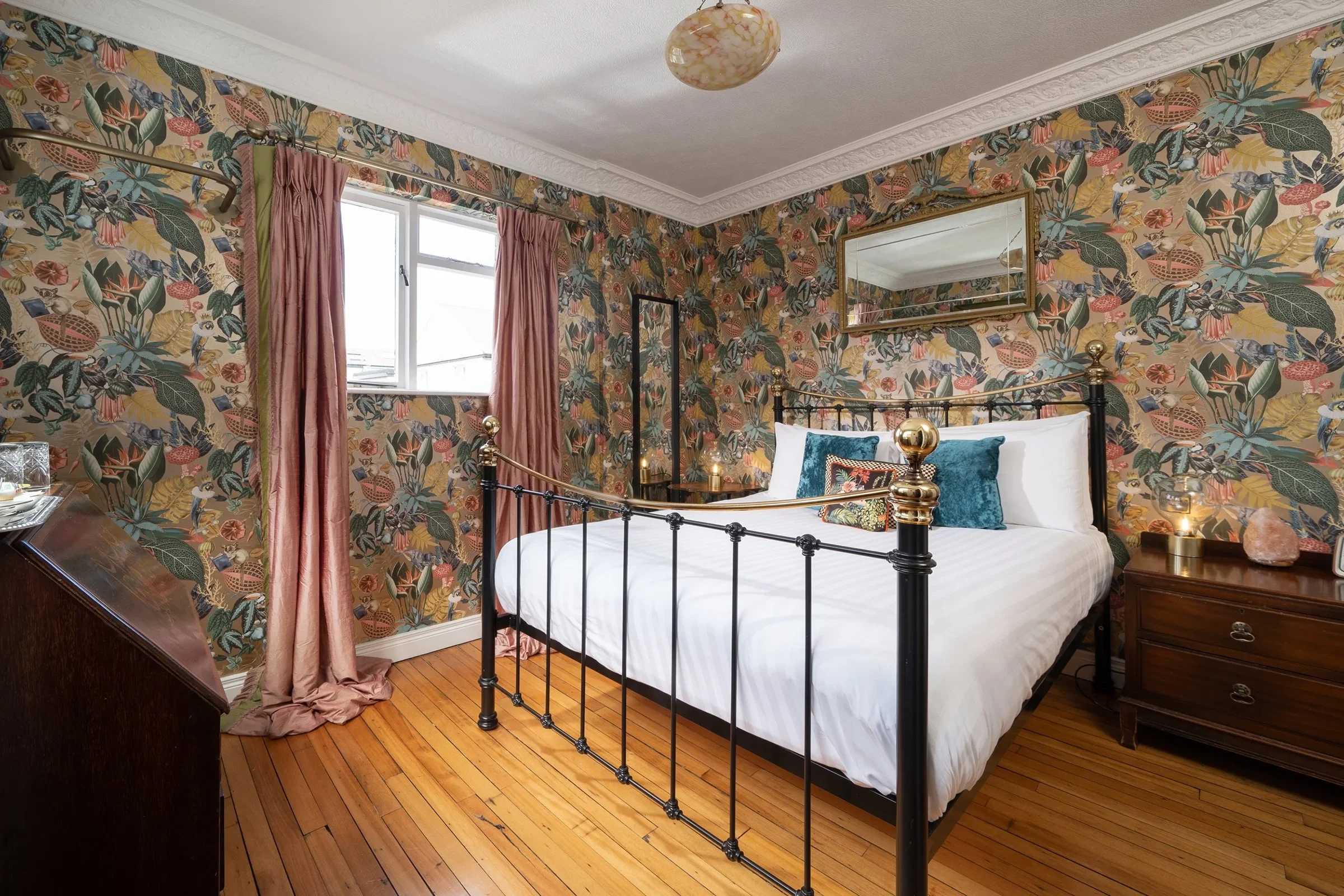 A bedroom with floral wallpaper, a brass and black metal bed with white bedding and decorative pillows, a wooden nightstand with a salt lamp, a mirror above the bed, pink curtains, and a window letting in natural light.